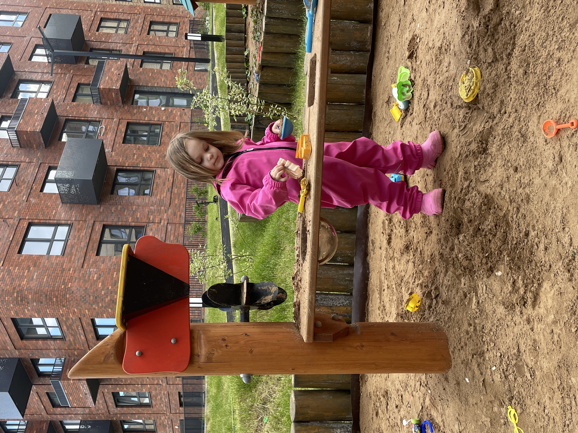 A young girl in a pink jumpsuit plays in a sandbox next to a wooden playground structure. Various plastic sandbox toys are scattered around the sand. Behind the playground is a modern brick apartment building with dark-framed windows and projecting black metal balconies. The scene is set outdoors with patches of grass visible between the sandbox and the building. The location is likely in Pervomaysky, Russia.