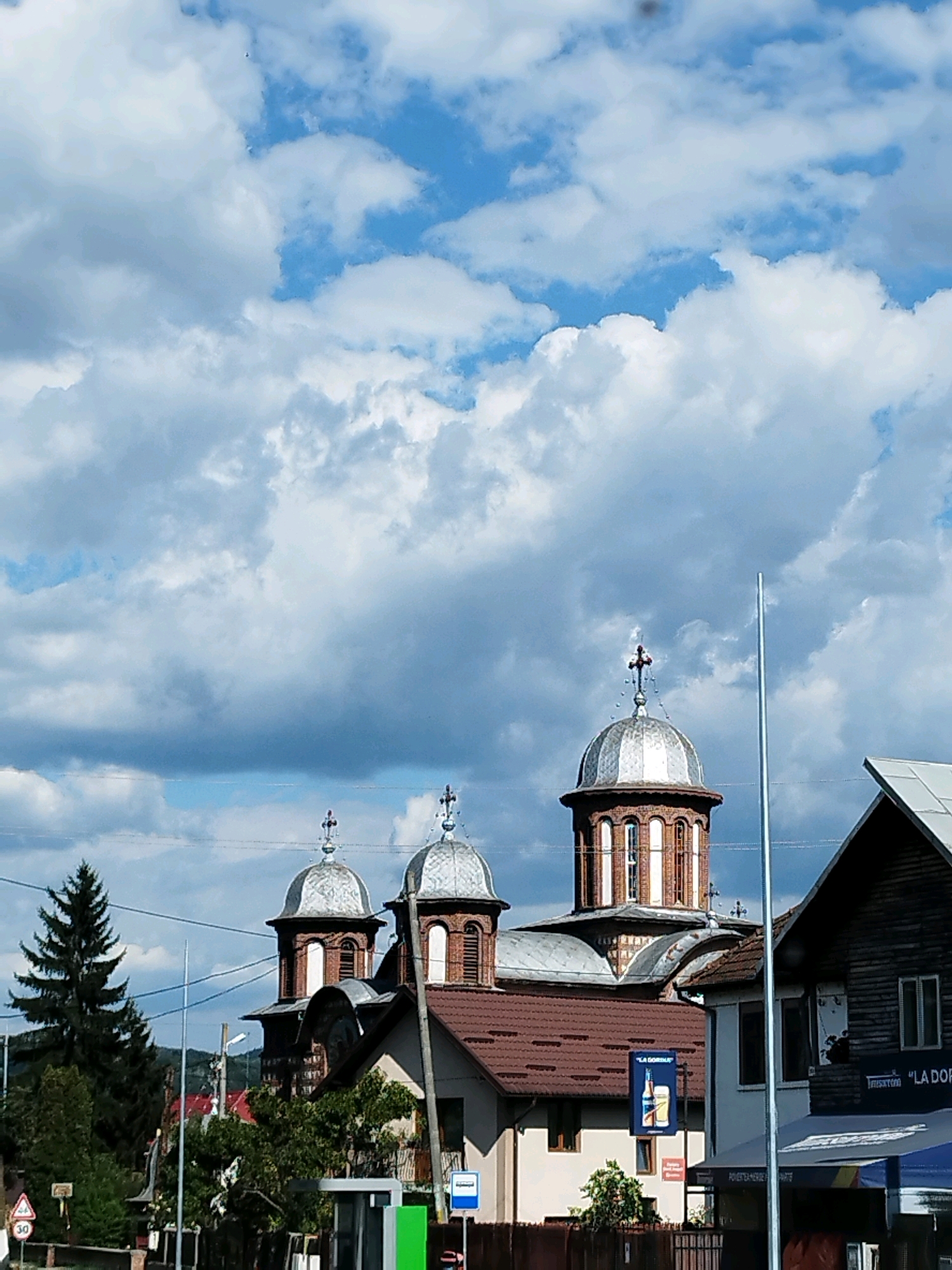The image depicts a view of the Curtea de Argeș Monastery in Curtea de Argeș, Romania. The monastery features a prominent copper-domed tower topped with a cross, characteristic of Orthodox Christian architecture. The main structure appears to be constructed from brick and stone. In the foreground, there are various buildings, including a wooden-clad structure and what seems to be a bus stop. A tall, slender metal pole stands to the right of the monastery. The sky is filled with dramatic clouds, with patches of blue sky visible. Trees, including a tall pine tree, are present to the left of the monastery. There are road signs and what appears to be a billboard advertising a beverage. The overall impression is of a typical street view in a Romanian town, with a significant religious landmark dominating the background.