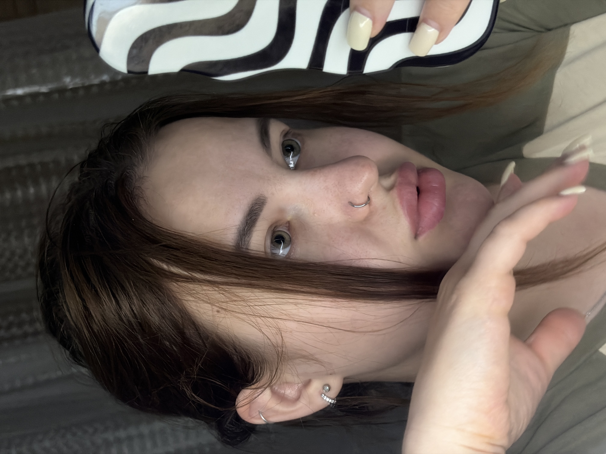 A young woman with light skin and brown hair poses with her face tilted toward the camera. She has a nose ring and multiple piercings on her ear. Her lips are full and pink, and she has light green eyes. One hand is near her face, the other is holding a white and black patterned object. The background consists of an olive-green fabric, likely a bed or couch. The location is not discernible from the image.