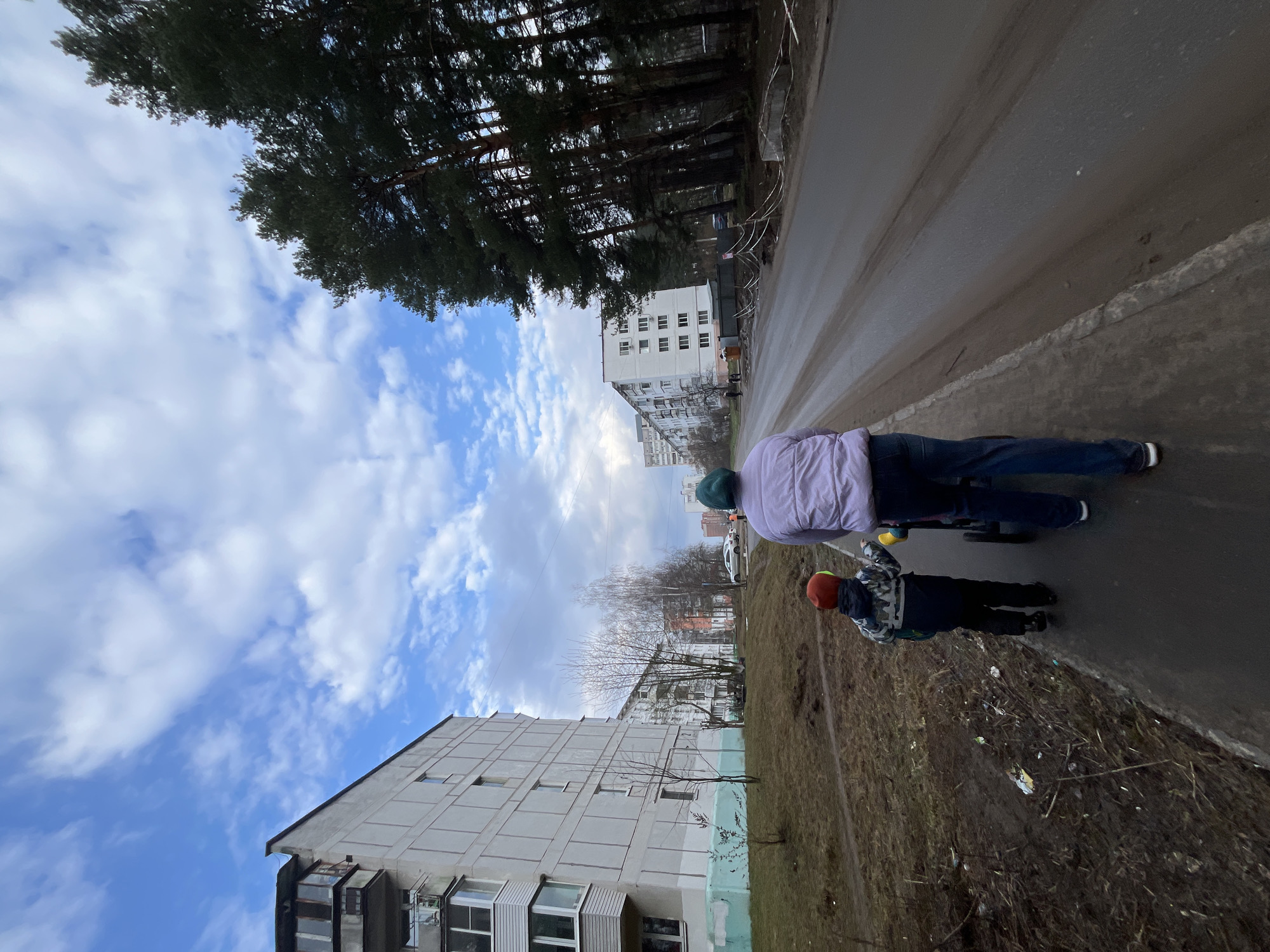 Two individuals are situated on a paved sidewalk next to a street. The taller person, wearing a light purple jacket, jeans, and white shoes, is riding a scooter. A younger individual, dressed in a dark jacket with a camouflage pattern and an orange helmet, is also on a scooter. The sidewalk is bordered by a patch of dry grass and debris. Across the street, there are multi-story apartment buildings, bare trees, and a dense forest in the background. The sky is partly cloudy. The location is in a residential area of Chaikovsky, Russia.