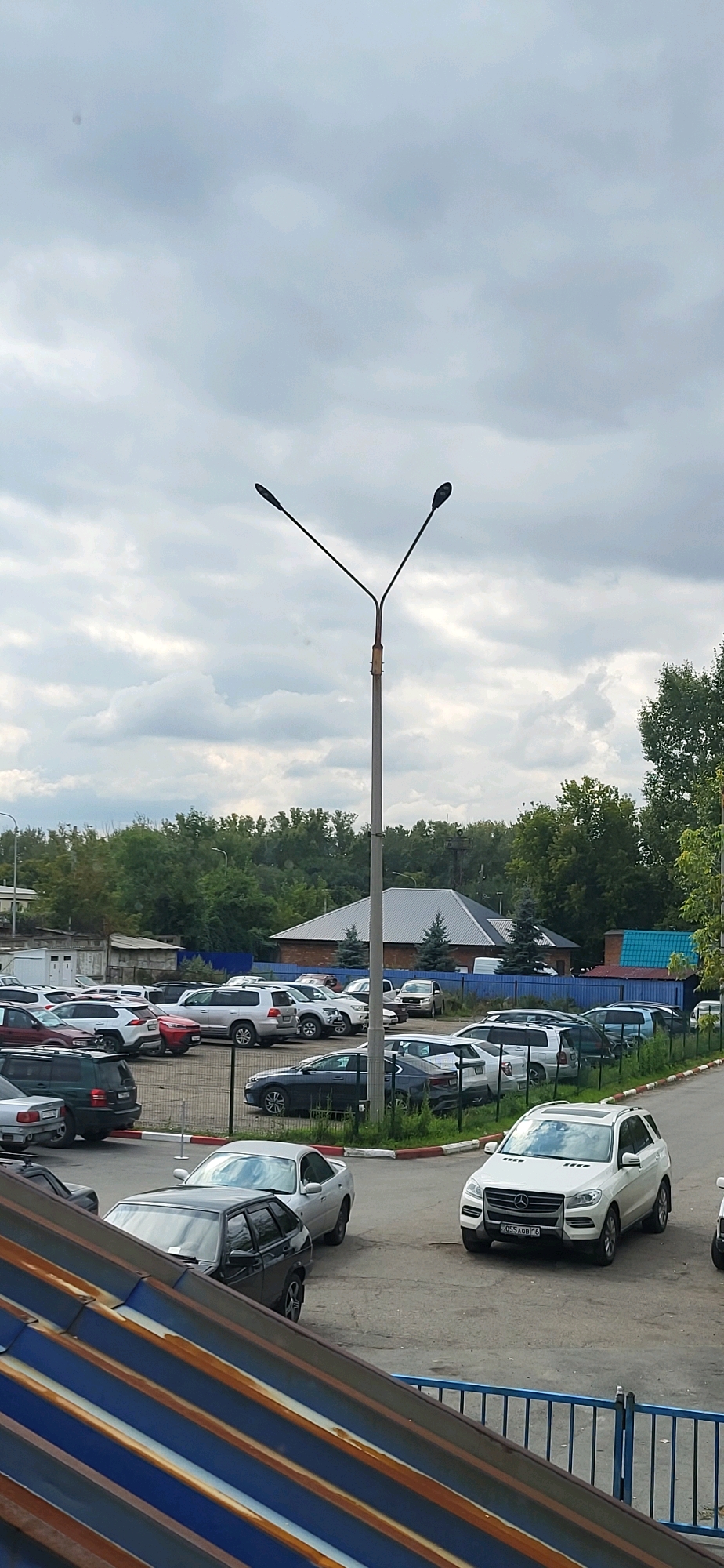 The image shows an outdoor parking lot on an overcast day. A tall, two-headed street lamp stands prominently in the center-right of the frame. The parking lot is filled with various vehicles, including sedans and SUVs. In the background, there are trees and buildings, one with a distinctive grey, angled roof. A blue fence runs along the back of the parking lot. The foreground features a glimpse of a slanted, multi-colored metallic roof structure and a blue railing. No people are visible in the scene. The scene is likely in the city of Oskemen, Kazakhstan.