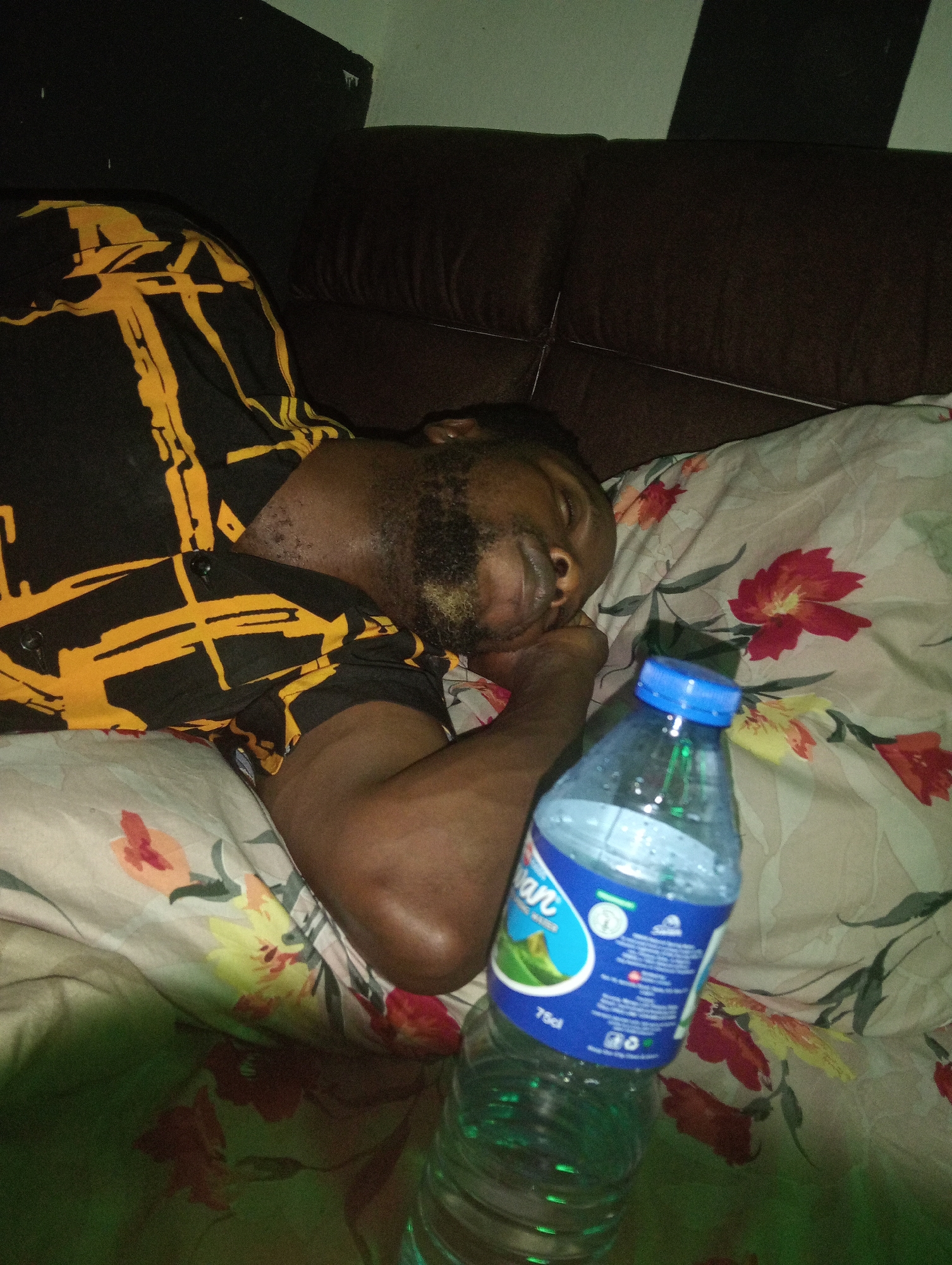 A bearded man is asleep on a flower-patterned pillow, resting his head on his right arm. He is wearing a black shirt with yellow rectangular patterns. A half-empty bottle of water with a blue cap is positioned in the foreground, near his hand. The setting includes a dark brown couch and the background suggests an interior space. The location, Jos, Nigeria, is given as requested but cannot be confirmed or denied from the visual data in the image.