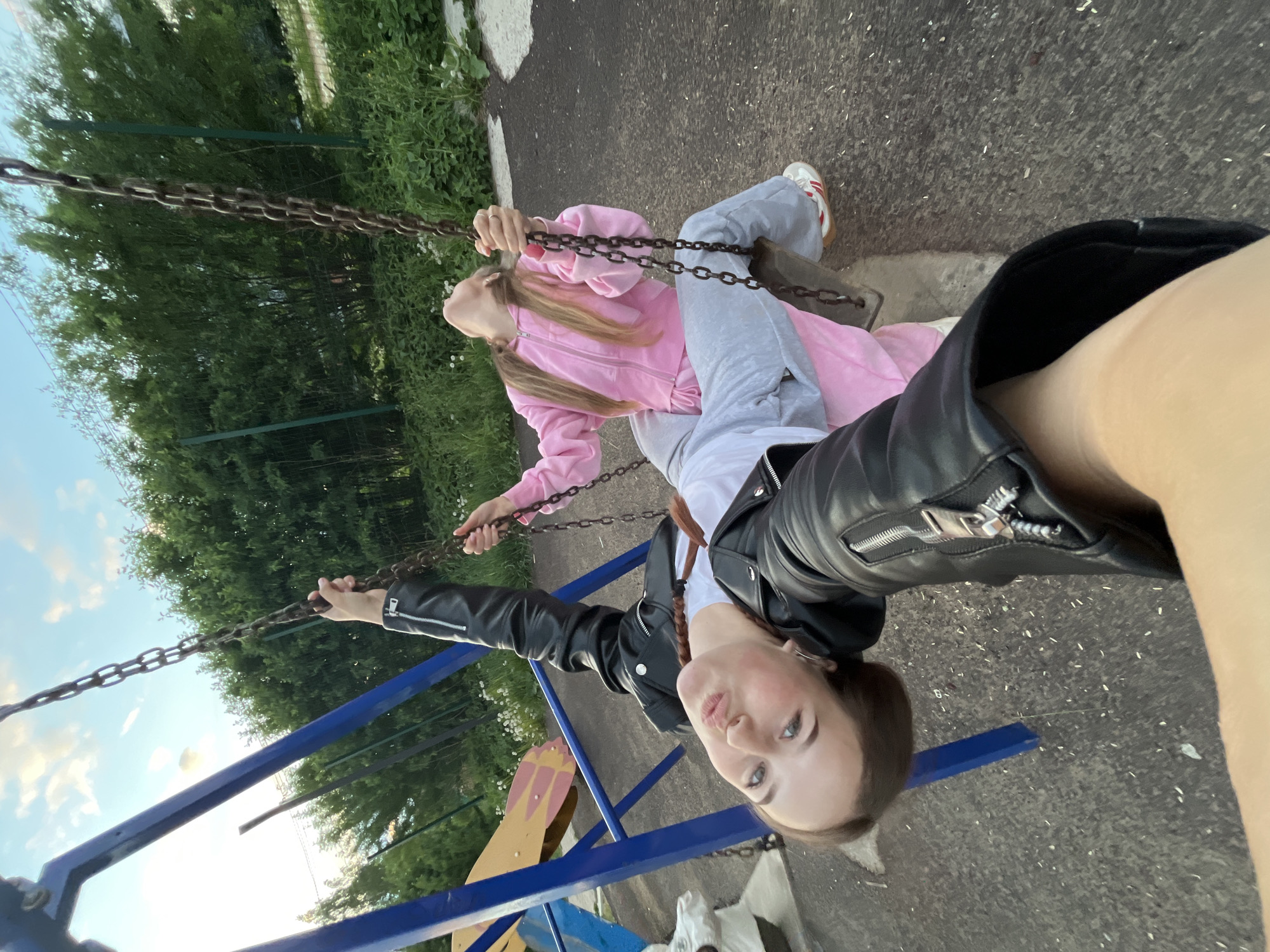 Two girls are on swings at a playground. One girl is wearing a pink tracksuit and is sitting on the swing. The other girl is wearing a black leather jacket and is standing on the swing while taking the photo. The playground is located in Фоки, Russia.