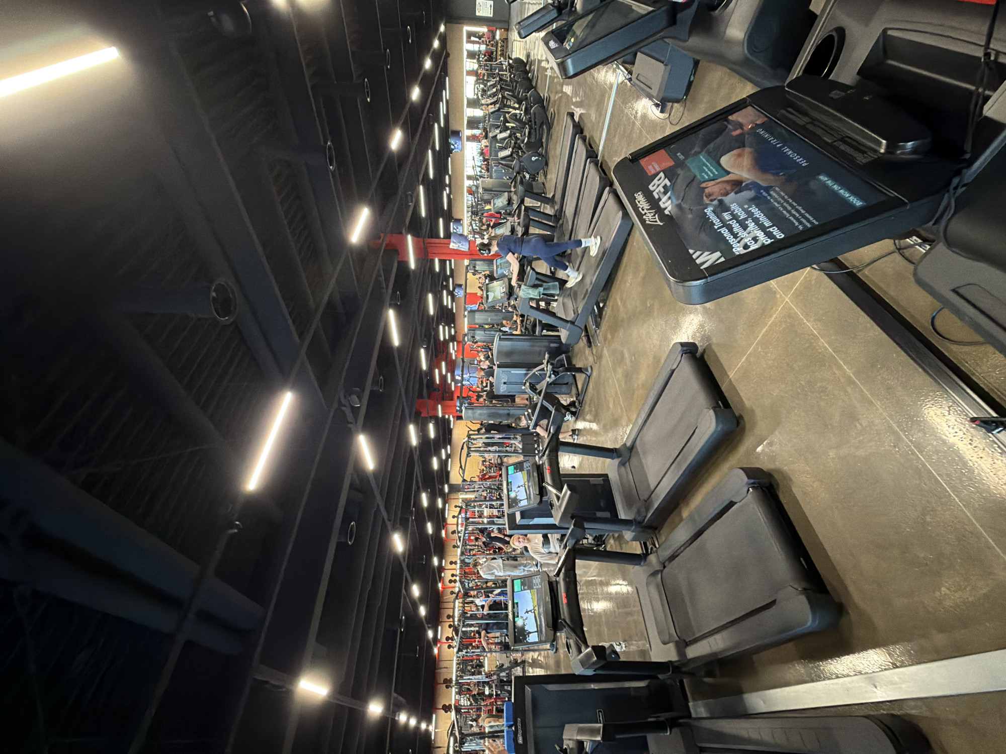 The image captures a gym interior featuring numerous treadmills arranged in rows. People are using the exercise equipment. The gym has an industrial aesthetic, characterized by dark, exposed ceilings, lighting fixtures, and visible ductwork. The location is likely Plovdiv, Bulgaria, due to the reference to "BG" in one of the OCR outputs.