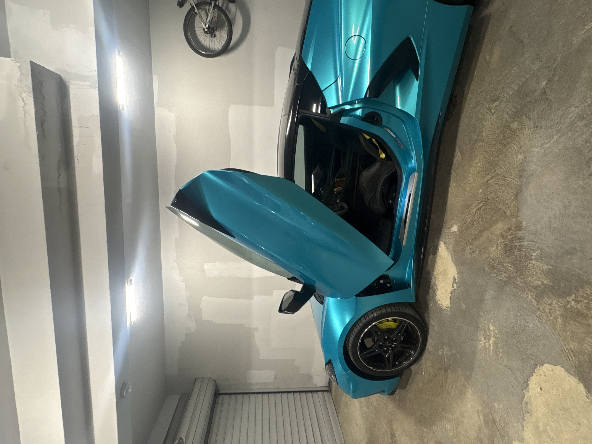 The image shows a turquoise sports car, likely a Corvette C8, parked in what appears to be a garage. The driver's side door is open, revealing a black interior with yellow accents. The car's wheel has black rims and yellow brake calipers. A bicycle hangs on the wall in the background. The garage has white walls, with a fluorescent light fixture visible on the left. The floor is concrete with visible wear and imperfections. There is no evidence to suggest that the image was taken in Varna, Bulgaria. No people are visible.