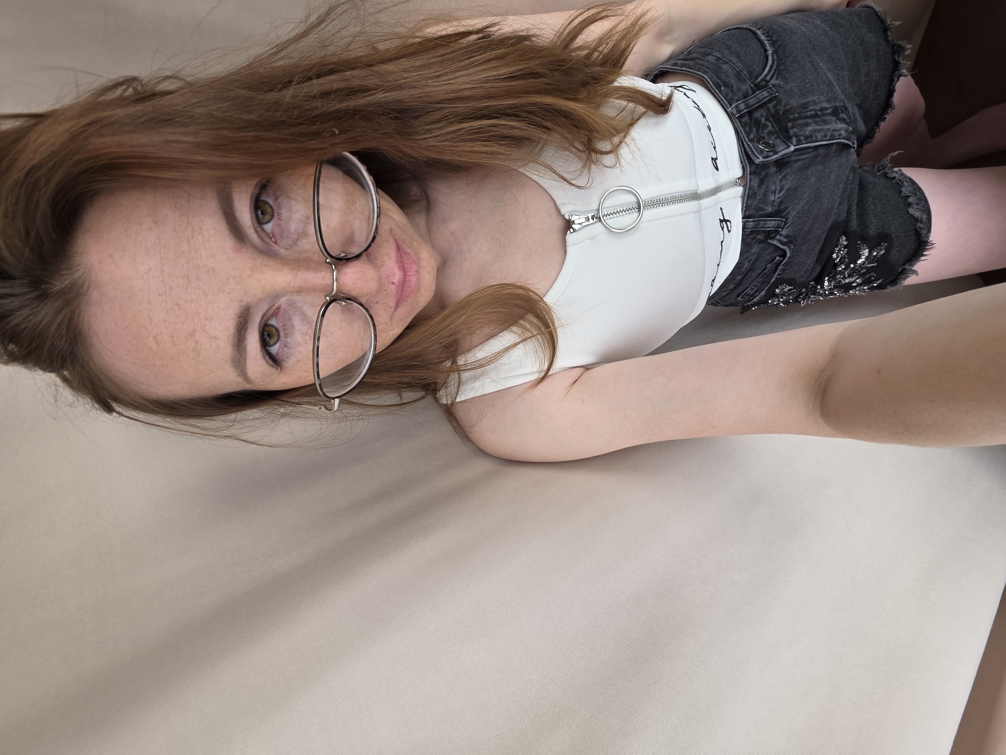A woman is lying on a beige surface. She has long reddish-brown hair, fair skin, and freckles. She is wearing round-frame glasses, a white zippered top, and dark denim shorts with some embellishments. The photo appears to be taken from above, at a close angle to the subject. The scene is not explicitly indicative of Chaikovsky, Russia.