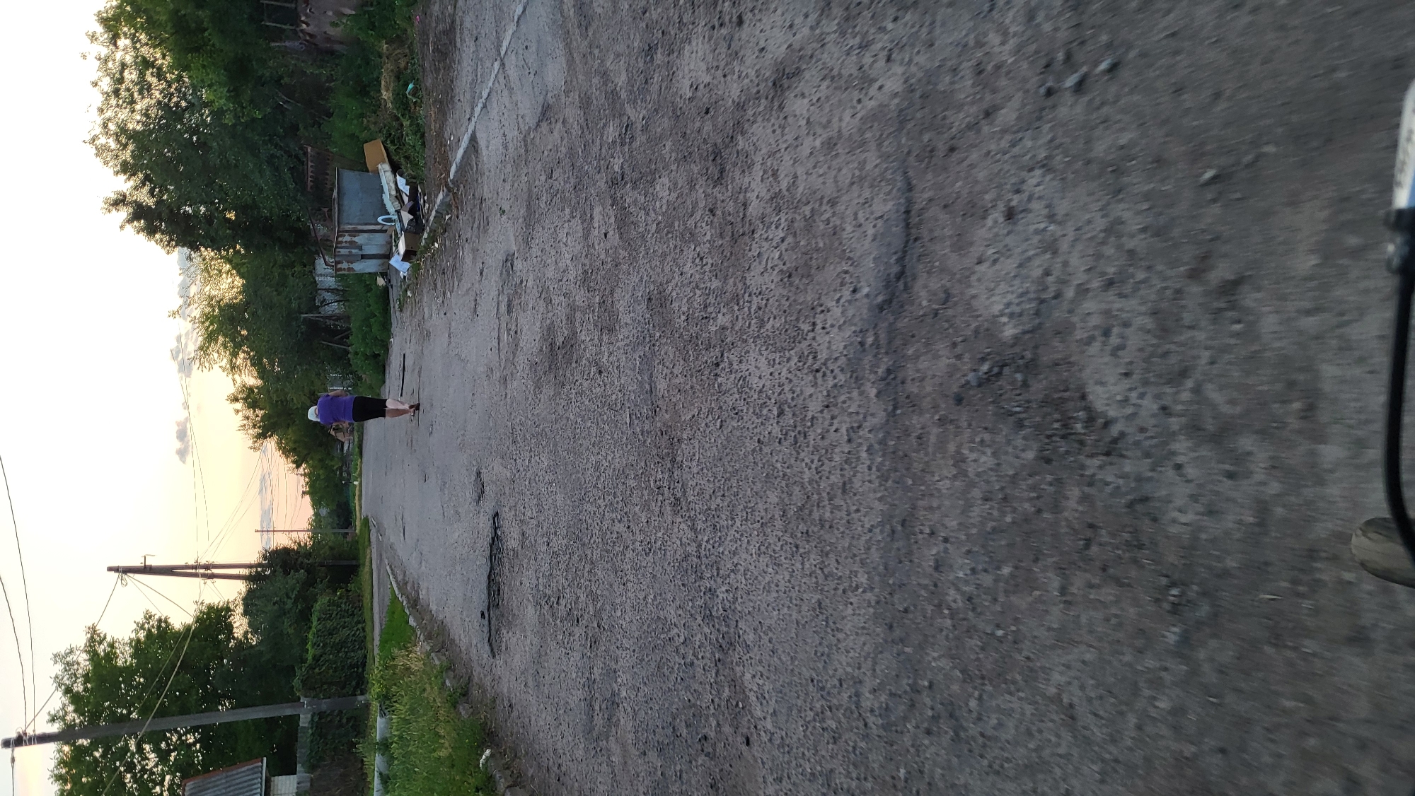 A person walks away from the camera on a cracked asphalt road in a rural or suburban area. The person is wearing a purple shirt and dark pants. The sky is a pale orange and white, suggesting either sunrise or sunset. Trees line the sides of the road, and there are some dilapidated structures and piles of debris visible. The road is in poor condition with several potholes. The perspective suggests the image was taken from a moving vehicle.