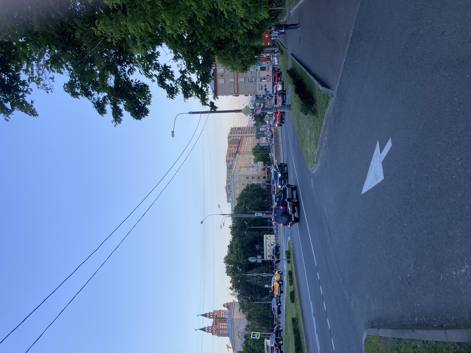 The image depicts a street scene in Khimki, Russia, likely during the late afternoon or early evening, as indicated by the soft light and clear sky. In the foreground, a dark asphalt road curves to the right, marked with a prominent white arrow indicating a right turn. To the left of the road, a strip of grass and a concrete curb delineate the edge of a green median with manicured bushes. Further left, across the street, is a multi-lane road with several vehicles, including a yellow taxi and a dark SUV.
In the middle ground, beyond the road, a row of trees forms a dense green barrier. Behind the trees, a large, ornate building with twin spired towers, possibly a cultural or governmental institution, is visible. Several streetlights are positioned along the road. On the right side of the image, a paved sidewalk runs parallel to the street, lined by a row of mature trees with lush green foliage. A few pedestrians are walking on the sidewalk, and a person wearing a backpack is walking away from the camera. In the background, apartment buildings and other urban structures are visible under a pale blue sky. The overall impression is of an active urban environment with a mix of residential and institutional architecture.