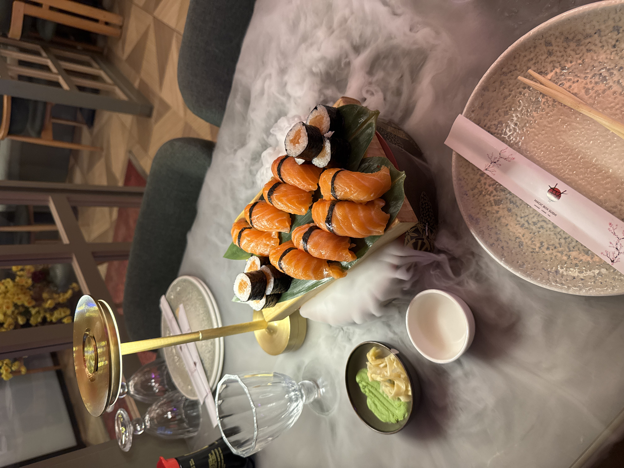 An overhead shot shows a sushi platter with several pieces of salmon nigiri and maki rolls, presented on a wooden board with a leaf garnish. The platter is surrounded by a cloud of dry ice mist, creating a dramatic effect. To the left of the platter are a small bowl of green wasabi and thinly sliced pickled ginger, a glass goblet, and a soy sauce bottle. A metallic lamp with a golden base and shade stands behind the platter. In the foreground, a plate with a decorative chopstick wrapper and a pair of wooden chopsticks rests on a table. The table surface is covered in a gray, textured tablecloth, and the background shows glimpses of a restaurant interior with tables, chairs, and wooden decorative screens.