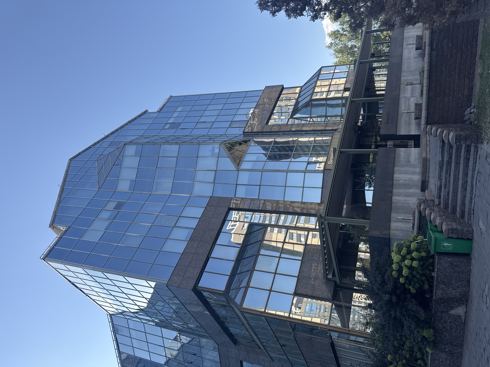 A low-angle, eye-level shot captures the exterior of a modern glass and granite building, likely a commercial or office space, under a clear blue sky. The building features a distinct geometric design with large, reflective glass panels forming most of its facade, interspersed with dark granite sections. The glass reflects the blue sky and other buildings, creating a dynamic visual effect.
At the base of the building, a covered walkway or patio area is visible, supported by thin columns. Lush green bushes and trees surround the lower levels of the building, with some autumnal foliage also present. Steps made of stone and brick lead up to an entrance. Two green garbage bins are situated near the bottom of the steps.
The building appears to be located in the city of Almaty, Kazakhstan, as indicated by the name "Benedict" visible on the facade in a subtle, dark font. The architectural style and the surrounding vegetation are consistent with urban environments in Almaty. No people are visible in the frame. The lighting suggests it is daytime, likely late morning or early afternoon, given the bright sunlight and clear sky.