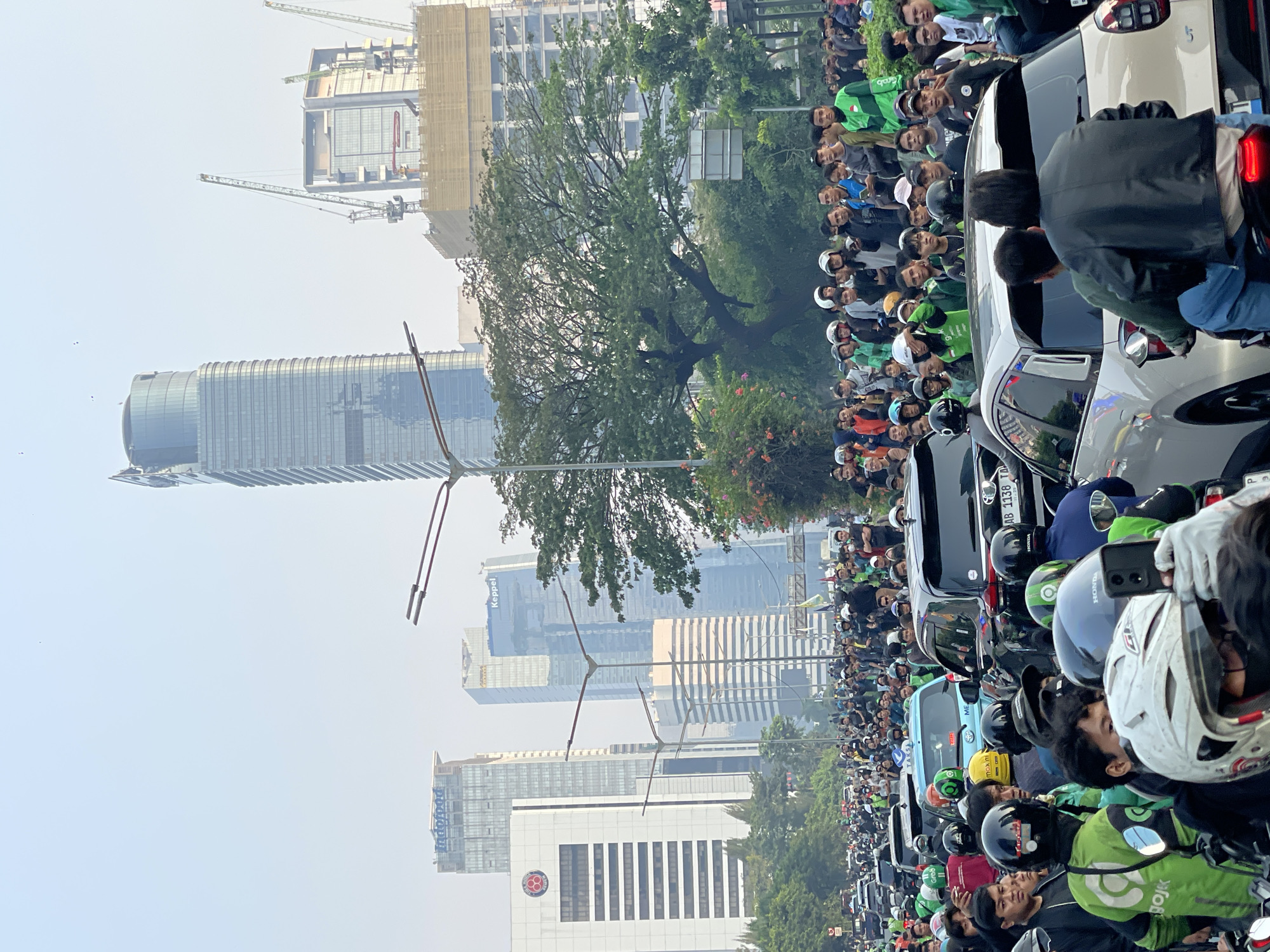 This is a high-angle, full shot of a busy street in Central Jakarta, Indonesia, on a hazy day. The scene is dominated by a large traffic jam of cars and motorcycles, with many people on foot gathered on the sides of the road. In the background, several modern skyscrapers pierce the sky, some of which are under construction, indicated by the presence of cranes. The buildings showcase a mix of glass and concrete facades, with prominent signage such as "Indofood" and "Keppel" visible on some structures.
The foreground and midground are filled with a multitude of individuals, many wearing green jackets and helmets, suggesting they are likely ride-sharing drivers. They are standing and milling about amongst the stationary vehicles. The traffic consists of various vehicles, including cars, SUVs, and numerous motorcycles. The atmosphere appears to be one of congestion and perhaps a gathering or protest due to the large number of people congregated. The sky is a pale, hazy blue, and the overall lighting suggests it is daytime, possibly late afternoon or early morning. The presence of trees and some flowering plants adds a touch of nature to the urban landscape.