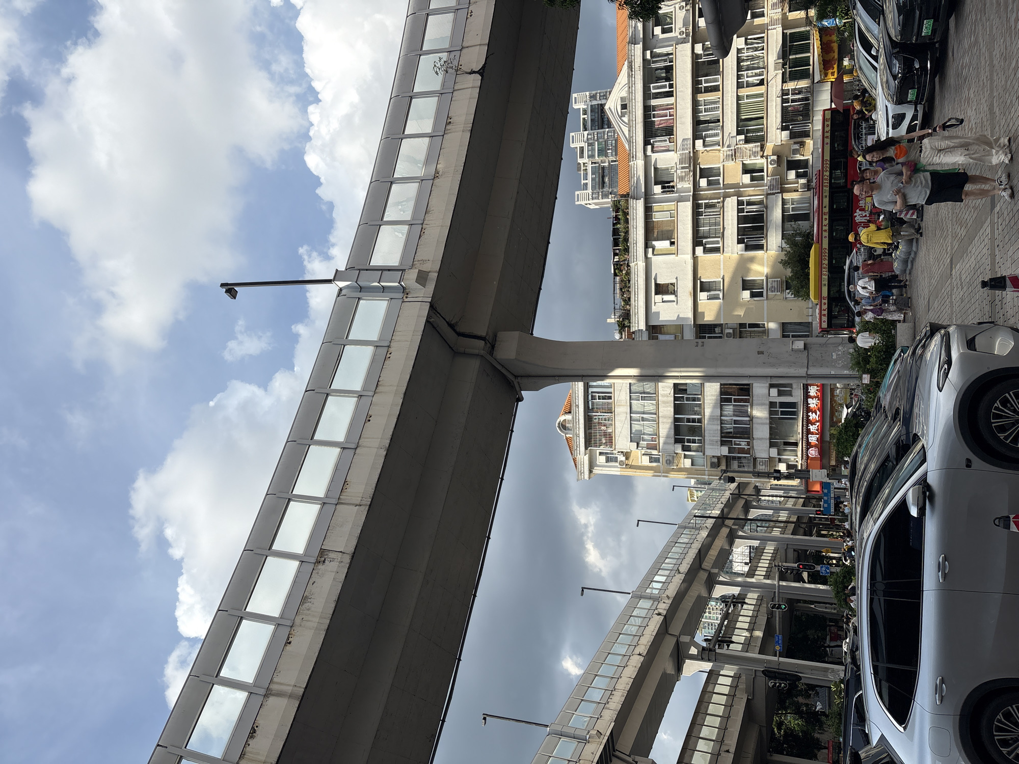 In Haizhu District, China, a street scene unfolds beneath a multi-level elevated highway system. The foreground features several parked cars, including a silver SUV and dark sedans. Beyond the cars, a group of pedestrians walks along the sidewalk, some carrying bags, while others appear to be conversing. A red and yellow bus with Chinese characters is visible further down the street. To the right, apartment buildings with numerous windows and balconies line the street, some displaying laundry or air conditioning units. The sky above is a mix of blue with scattered clouds and darker, overcast areas, suggesting dynamic weather conditions. The elevated highway structures are concrete, with transparent barriers along their edges. Streetlights are present on poles along the highway and on the ground level. The overall impression is a bustling urban environment with significant infrastructure.