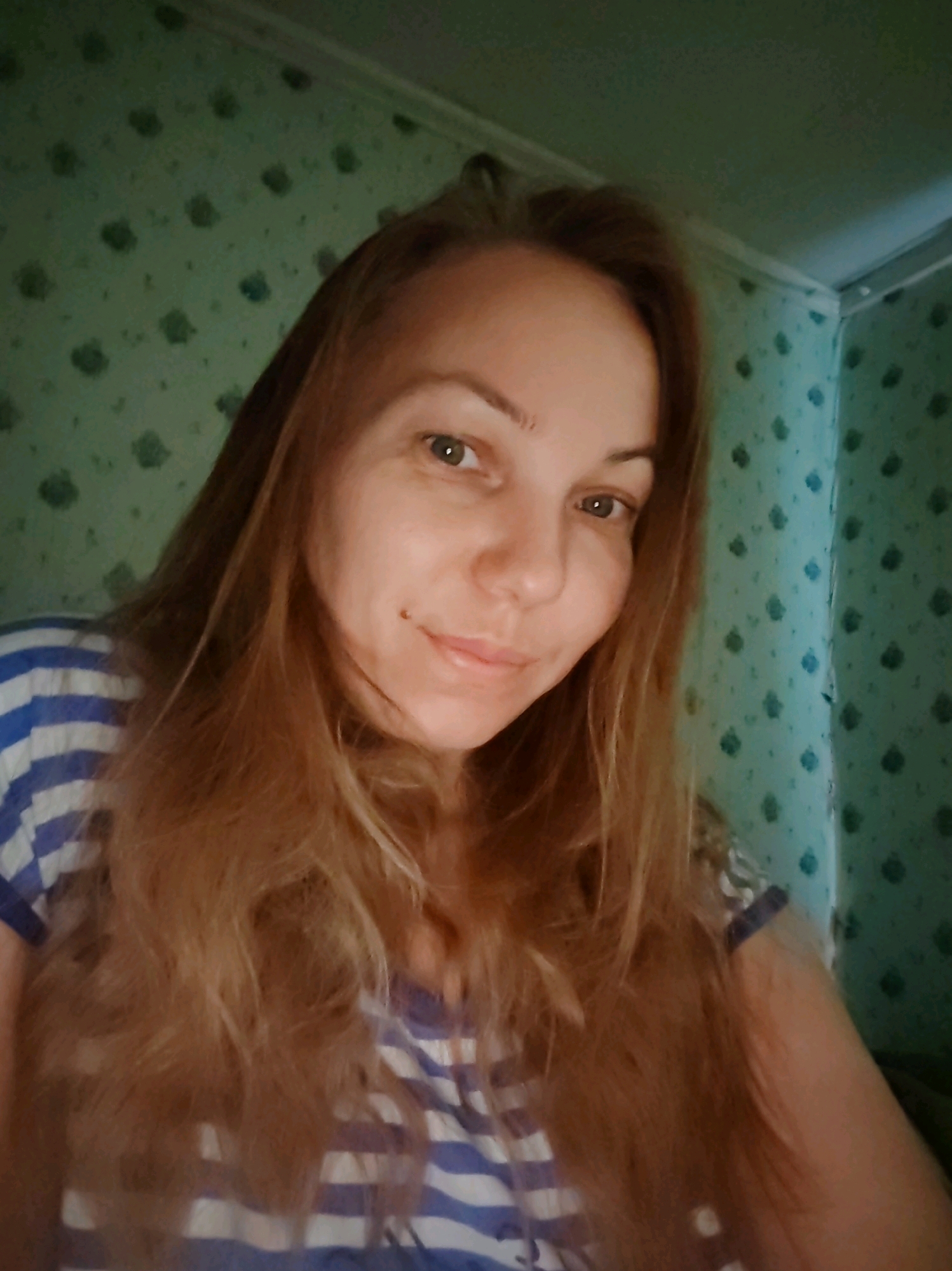 A young woman with long, wavy blonde hair and light green eyes is captured in a close-up, high-angle selfie. She is wearing a blue and white striped t-shirt. Her face is turned slightly to the right, and she has a mole on her left cheek. The background consists of light green wallpaper with a subtle floral pattern. The lighting suggests an indoor setting, possibly a room in an apartment or house, with the light source appearing to be from above and slightly to the front. The overall impression is casual and personal. The location is specified as Chuchlem, Russia, though the image itself does not provide any explicit visual clues to confirm this location.