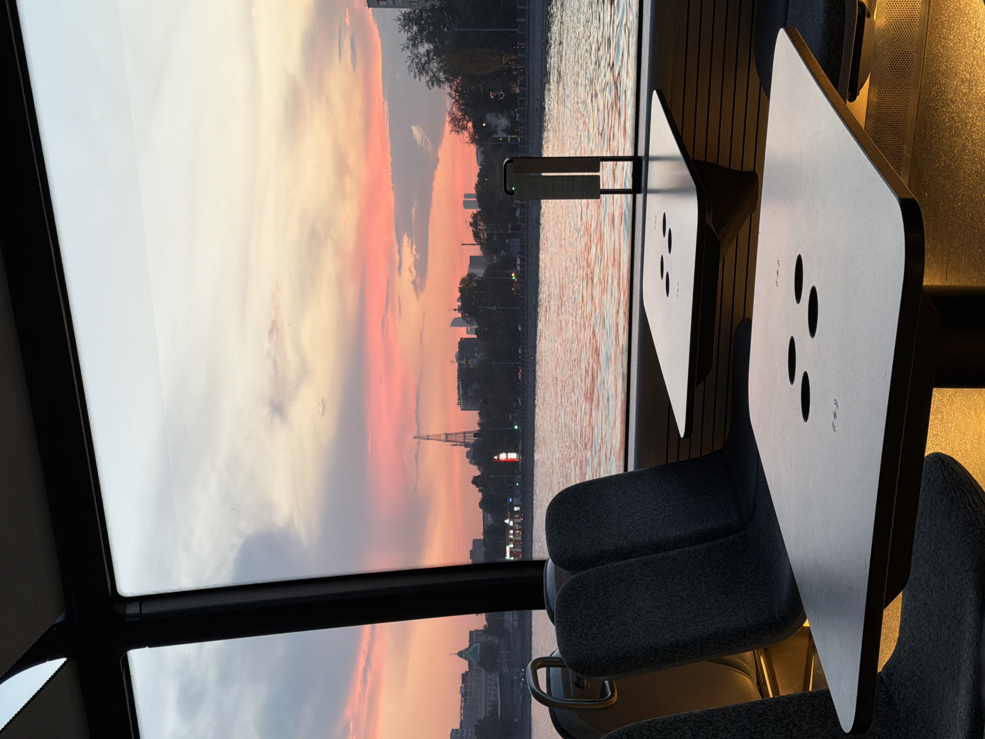 The image captures an interior view from a moving vessel on a river, looking out at a city skyline during sunset. Two tables with minimalist designs and integrated circular cutouts are prominently featured in the foreground, positioned on either side of dark upholstered seating. The vessel's interior is dimly lit, with warm yellow light emanating from beneath the tables, creating a cozy atmosphere.
Through the large windows, a body of water reflects the vibrant colors of the twilight sky, with hues of pink, orange, and purple dominating the scene. The Moscow skyline is visible in the distance, characterized by a collection of buildings, trees, and architectural landmarks, including a distinctive tower that appears to be the Ostankino Tower. The riverfront features a promenade and buildings, suggesting an urban setting within Moscow, Russia. The overall impression is one of a tranquil evening cruise with a picturesque view.