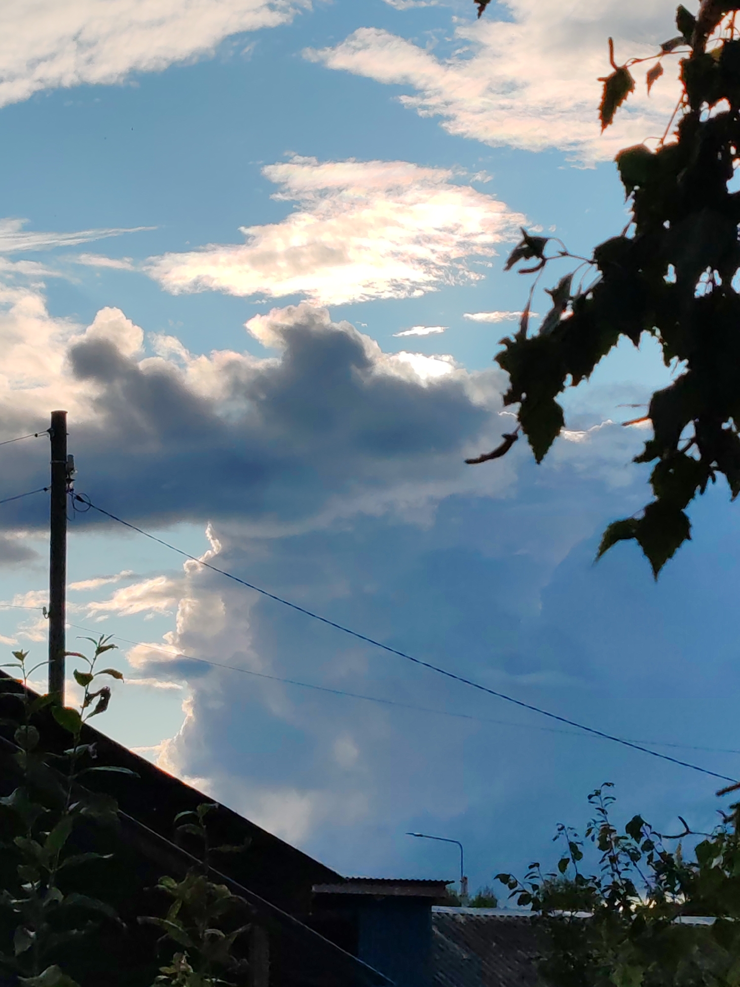 The image depicts a cloudy sky with a sliver of sunlight breaking through. In the foreground, partially silhouetted against the sky, are dark green leaves and branches of a tree on the right, and on the left, a dark rooftop with some foliage. A utility pole with wires runs vertically on the left side of the frame, crossing a diagonal wire. The sky is a mix of deep blue, gray, and white clouds, with some parts illuminated by the sun. The overall impression is of an overcast or partly cloudy day with the sun attempting to emerge. The location is stated to be in the city of Chukhlem, Russia.