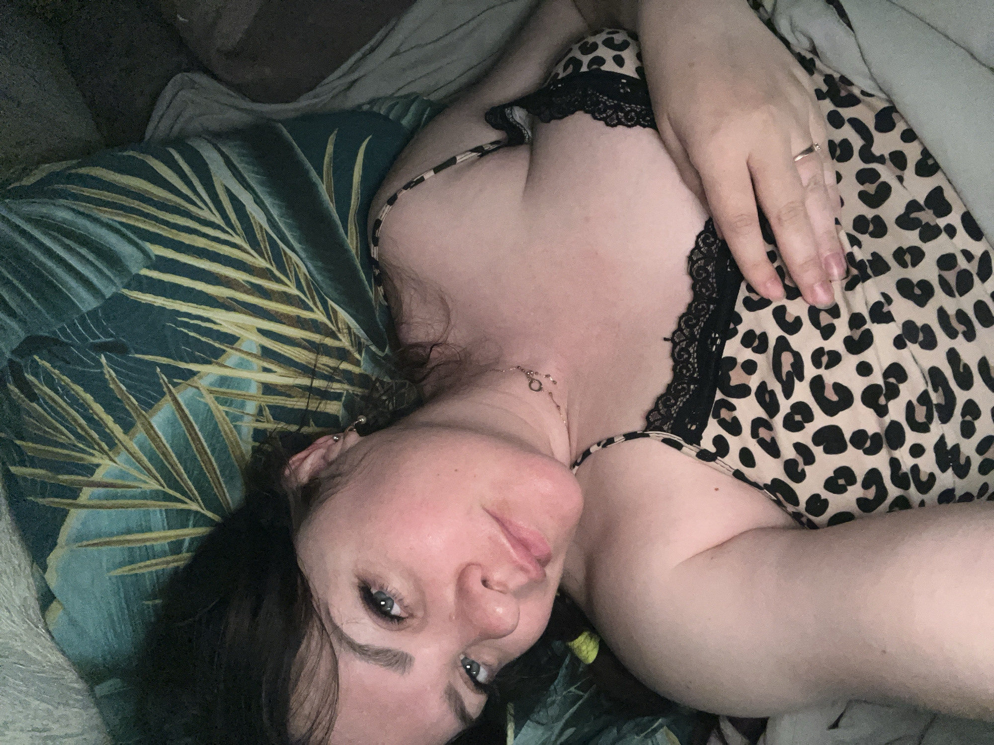 A woman is lying down, facing the camera. She has fair skin, dark hair, and blue eyes. She is wearing a leopard print top with black lace trim. Her left hand is resting on her chest, and she is wearing a ring on her ring finger. She is lying on a bed with a tropical print pillow. The location is unknown.