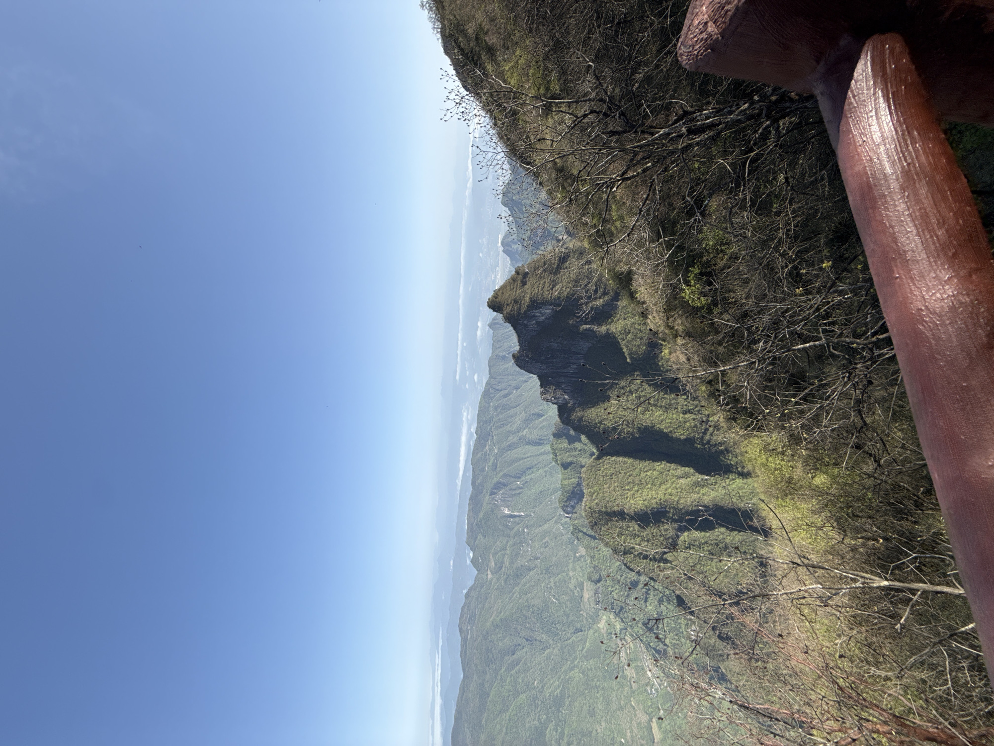 This image presents a stunning, expansive view of a mountainous landscape, likely captured from a high vantage point in Yongding District, China. The scene is dominated by rugged, verdant mountains and steep cliffs that recede into the distance under a clear, bright blue sky.
In the foreground, on the right side, a sturdy, reddish-brown railing or protective barrier is visible, suggesting the photograph was taken from a designated lookout point or path along the cliff edge. Bare tree branches, some with small buds or dried leaves, are prominent in the foreground and along the immediate cliff face, hinting at a season of dormancy or early growth.
The mountains are covered in a varied tapestry of dense green foliage, with some areas appearing darker or rockier. The distant peaks are softened by atmospheric haze, creating a sense of depth and vastness. The bright, clear blue sky occupies a significant portion of the upper frame, indicating excellent weather conditions. The overall lighting suggests a sunny day, providing clear visibility of the intricate geological formations.
No specific activities or events are depicted, the focus remains purely on the natural grandeur of the mountain range. No text is visible in the image.