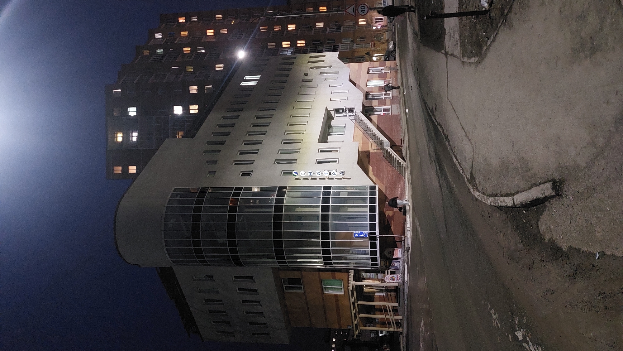 The image shows a street scene in Novosibirsk, Russia, at night. A multi-story building with a curved facade dominates the view, and its name "СОМПА" is visible. The building has a large glass-fronted section on the lower floors. Adjacent to this building is a taller, more conventional residential building with numerous illuminated windows. A person is standing on the sidewalk near the building. A street with a curb runs along the right side of the image. There is street lighting, and the sky is dark.