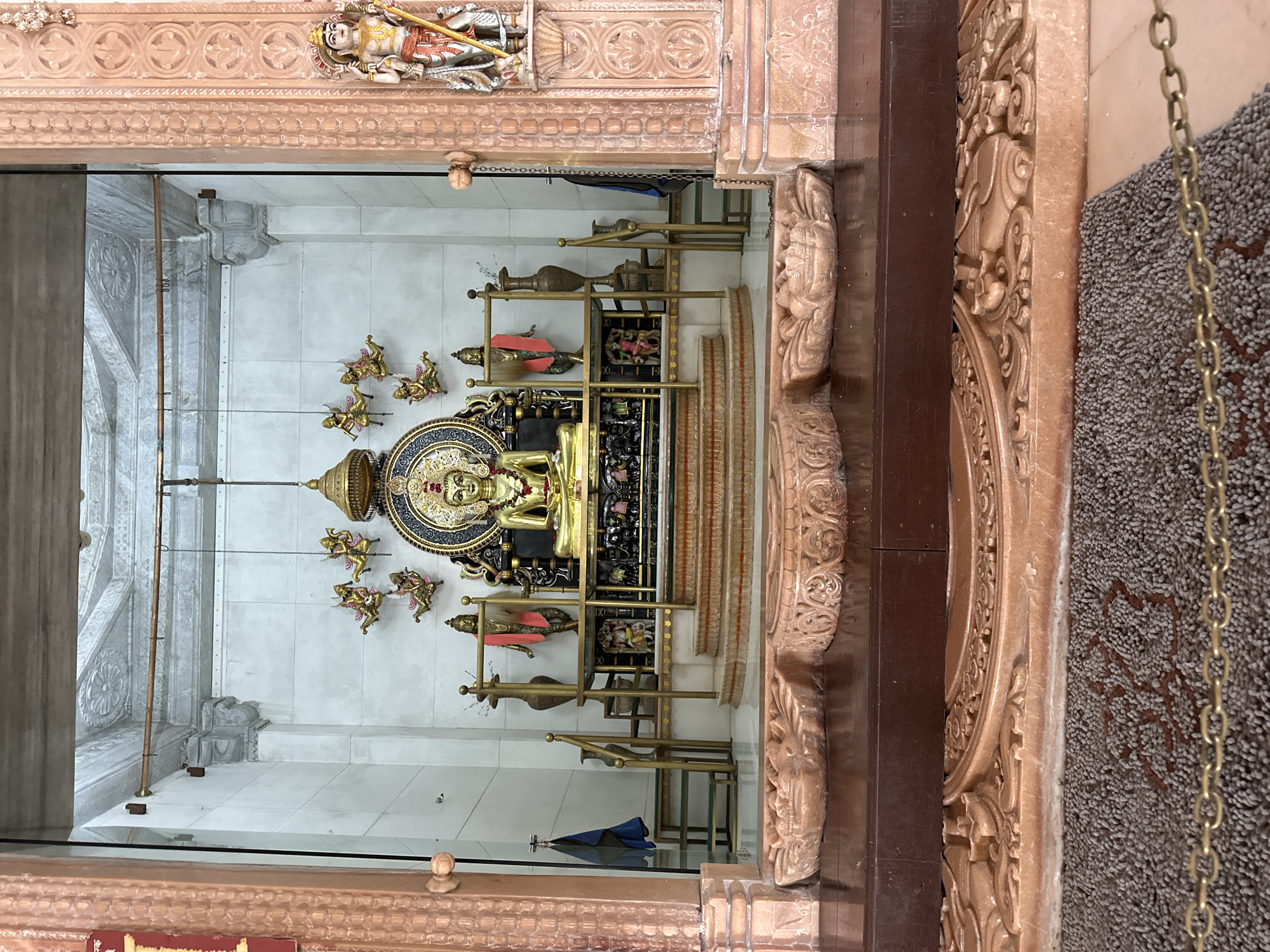 The image shows an interior view of a religious shrine, likely a temple, with a golden statue of a seated figure prominently displayed on a raised platform. The statue is ornate, with a halo and intricate detailing. The platform is tiered and accessible by steps. There are also smaller statues and decorative elements around the main figure, including what appear to be flying figures suspended from the ceiling. The shrine features ornate pillars on the right and left sides, adorned with carvings. The overall impression is one of reverence and artistry. The description mentions Shahapur, India, suggesting this is a specific location.
