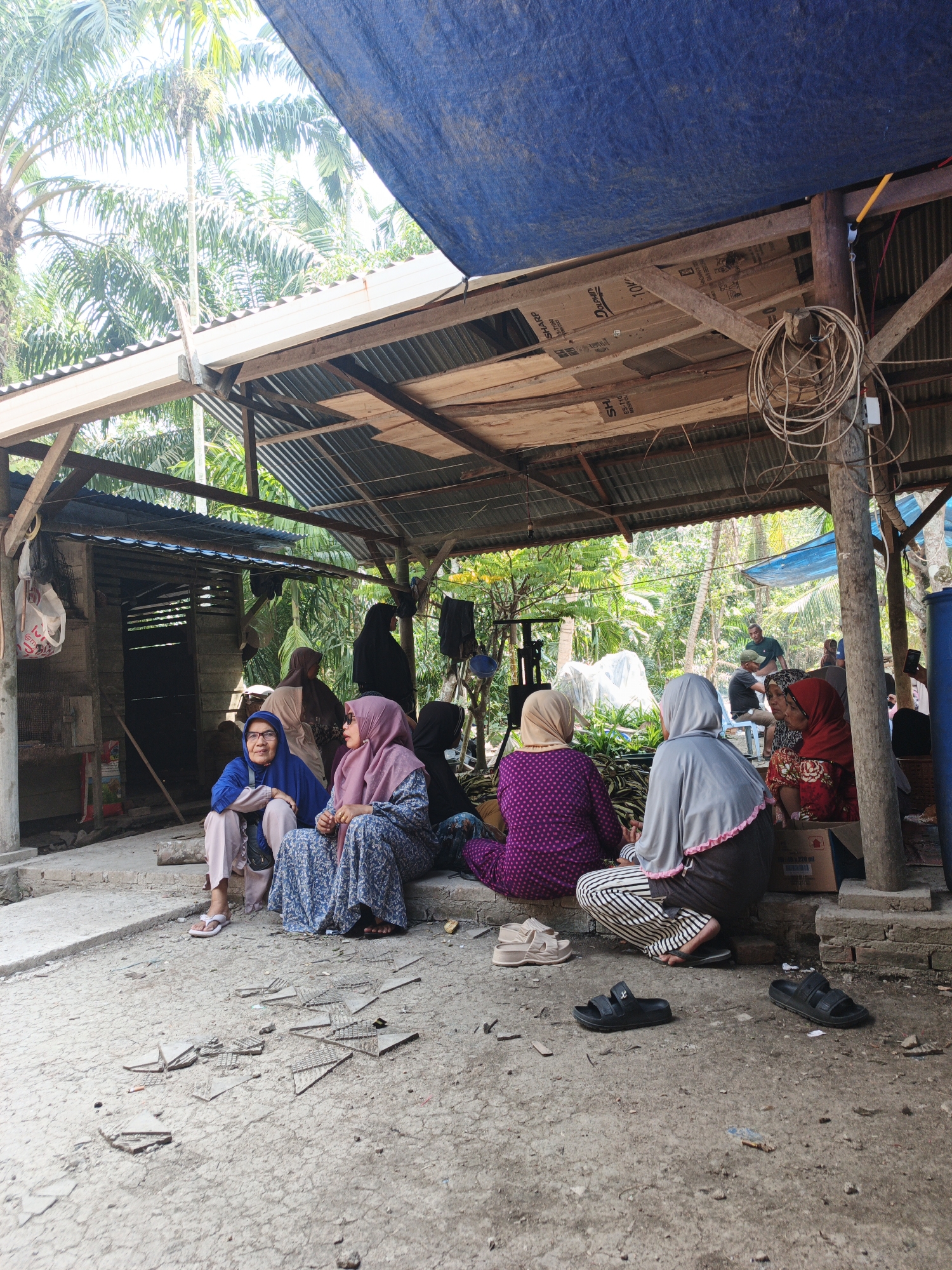 In the village of Pematang Duku, Indonesia, a group of women are gathered under a rustic, open-air shelter constructed with corrugated metal roofing supported by wooden beams and posts. The shelter is partially covered by a blue tarp, offering shade from the tropical sun. The ground is a mix of dirt and concrete, with scattered debris.
Several women, all wearing hijabs, are seated on the ground in conversation or observing their surroundings. In the foreground, two women sit close together; one in a blue hijab and light-colored pants, the other in a pink hijab and a patterned blue dress. To their right, another woman in a grey hijab and striped pants is crouched. Further back, other women are visible, some in darker attire.
The environment is lush and green, with palm trees and dense foliage visible behind the shelter, indicating a rural or semi-rural setting. An open wooden structure, possibly a shed or building, is visible to the left. Various objects are scattered around, including shoes, a coiled rope hanging from a post, and some boxes. The overall scene suggests a gathering or community event taking place outdoors.