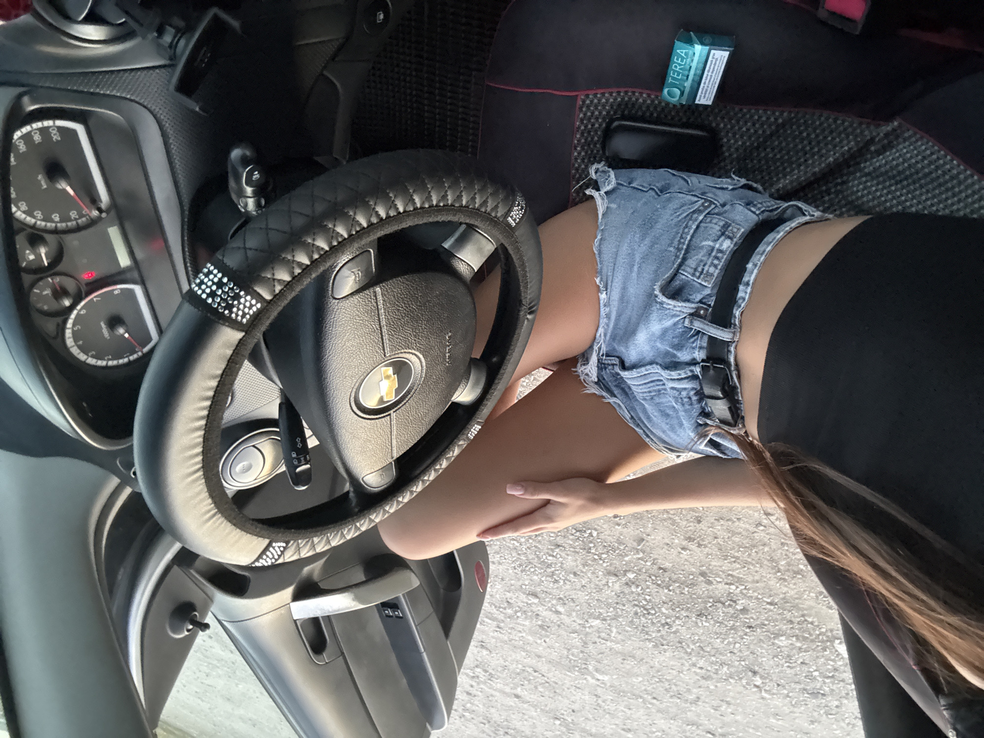 A person is seated in the driver's seat of a vehicle, outdoors in what appears to be Cimișlia, Moldova. The person, wearing a black crop top and denim shorts, has their right hand resting on their thigh. Their left arm is partially visible, extended towards the steering wheel. The interior of the car shows a black steering wheel with a chrome emblem, a dashboard with multiple gauges, and a car door. A small, rectangular blue box with white lettering is placed on the floor mat. The ground outside the vehicle is covered with gravel.
