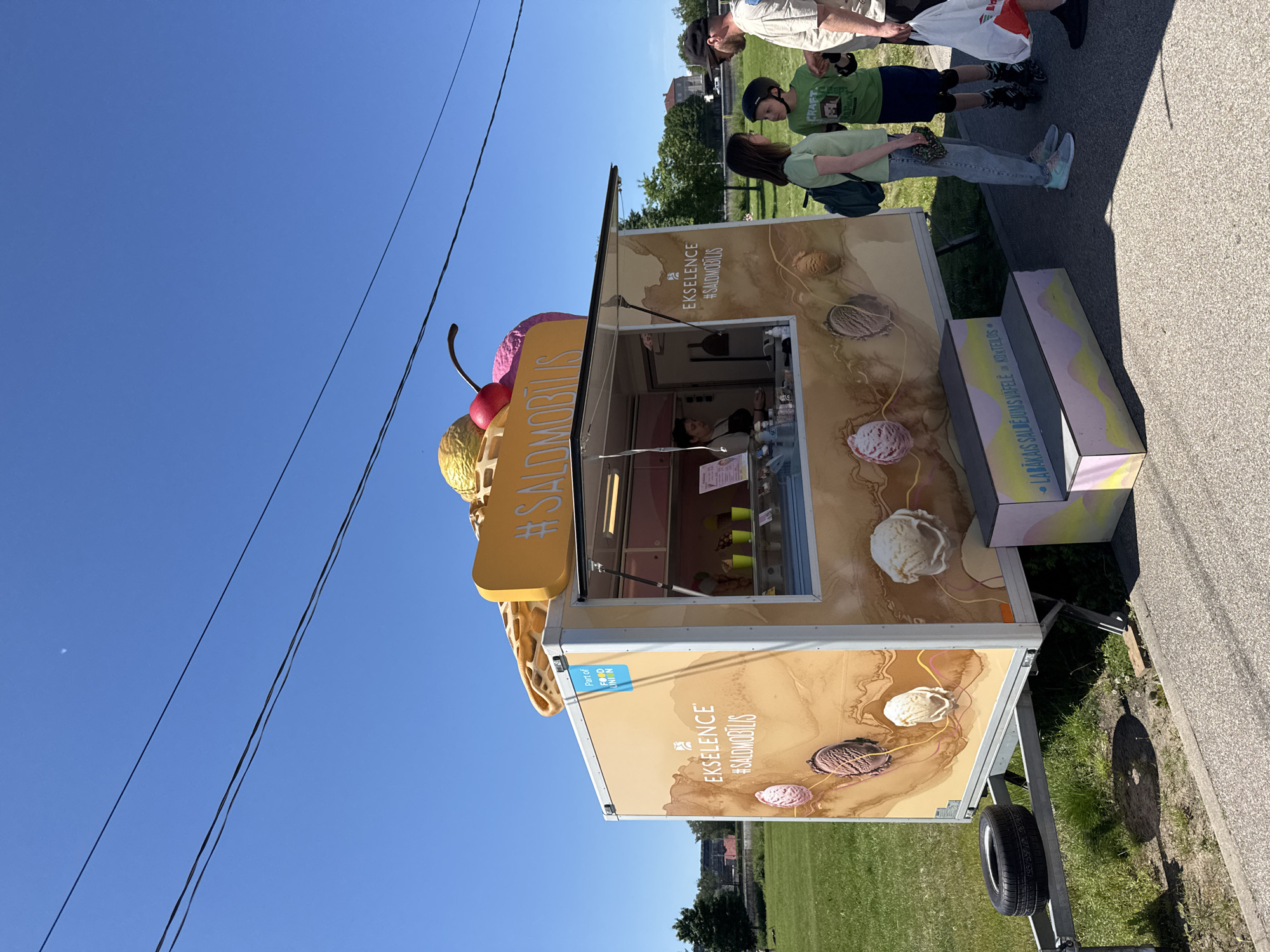 The image displays an outdoor scene featuring a mobile ice cream vendor, identified as an "EKSELENCE #SALDMOBILIS" trailer, on a clear, sunny day. The branding, which includes "Part of FOOD UNION" and Latvian text, indicates the location is in Latvia, likely in Riga given the urban context.
The trailer is light beige with large graphics depicting various scoops of ice cream (strawberry, chocolate, vanilla) against a swirling brown background. On its roof, a large 3D model of a waffle cone topped with yellow and pink ice cream scoops and a red cherry is visible. An open serving window is present on the right side of the trailer, where a female vendor in a dark apron is visible inside, with green cups on a counter. Below the serving window, two multi-level steps, painted in pastel pink, yellow, and blue, display Latvian text that translates to "BEST ICE CREAM WAFFLE OR COCKTAIL."
To the right of the trailer, three individuals are standing on a paved path. A woman with dark hair, wearing a light green t-shirt, blue jeans, and a dark backpack, stands facing the trailer. Beside her is a boy, approximately 8-10 years old, wearing a green t-shirt with "CRAFT" and "REPEAT" text, dark shorts, elbow pads, a black helmet, and rollerblades. He is looking towards a man positioned to his right. This man, wearing a light t-shirt and a dark baseball cap, holds a white plastic bag with red lettering in his left hand.
The immediate foreground consists of the paved path. Behind the trailer and individuals is an expansive green grass field with scattered trees in the mid-ground. Distant buildings, including one with a red roof, are visible on the horizon beyond the trees. The sky is clear blue, traversed by multiple black power lines in the upper portion of the frame. A faint, small, circular white object, possibly the moon, is present in the upper left quadrant of the sky. A spare car tire rests on the metal hitch frame of the trailer on the far left. Strong shadows are cast on the ground, indicating direct sunlight.