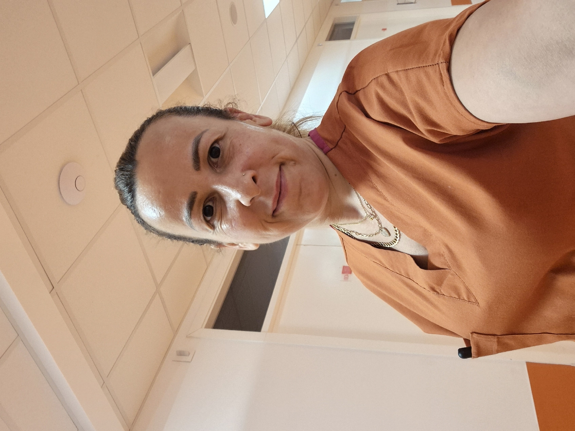 A woman with her hair pulled back in a bun, wearing an orange V-neck scrub top and two gold necklaces, poses for a selfie in a hallway. A black pen is tucked into the pocket of her scrub top. The hallway has white walls, a tiled ceiling with fluorescent lighting, and an orange accent color on the lower portion of the walls. The woman has a nose ring and is smiling at the camera.