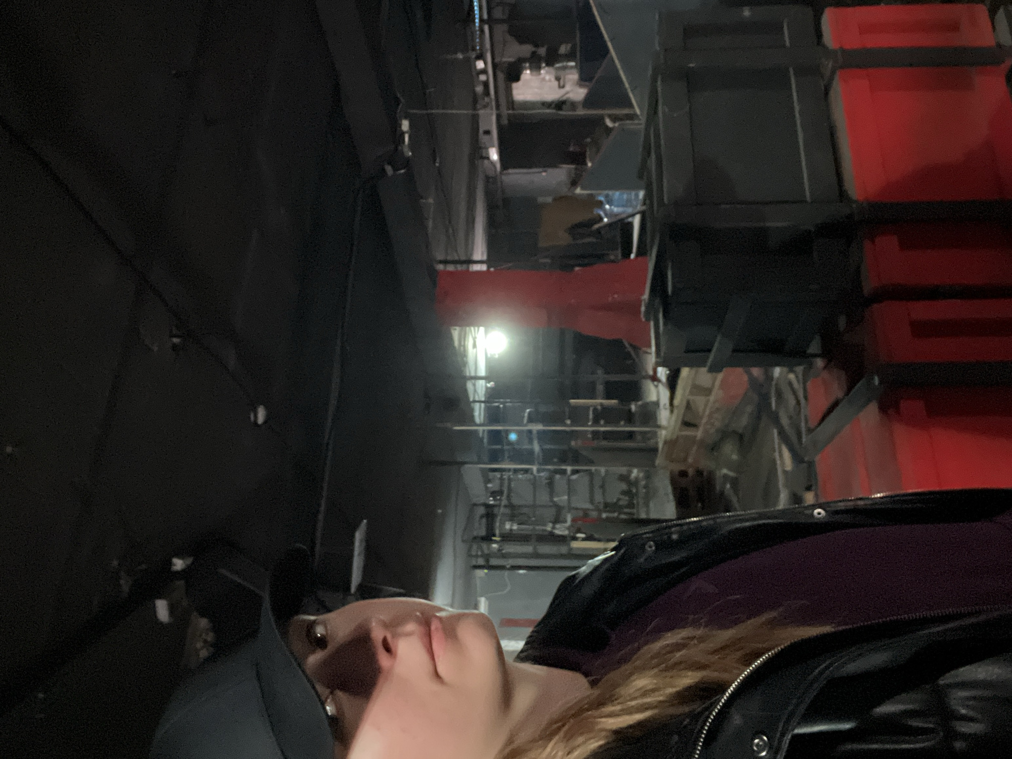 A person is looking up at an industrial-looking space. The person is wearing a black cap and a black jacket over a purple shirt. The industrial space above has metal beams and supports, a bright light source, and red containers. I cannot determine the specific location in Mytishchi, Russia.