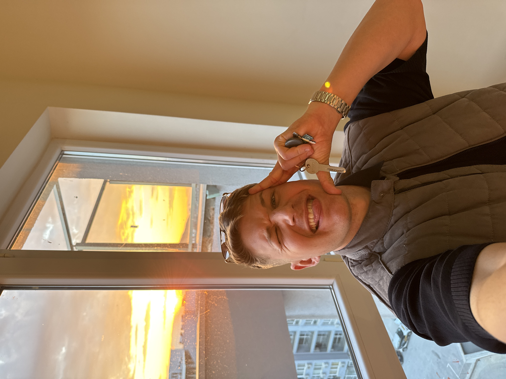 A person is holding keys to their head while standing by a window. Reflected in the window is an adjacent building and an orange sky suggesting either a sunset or sunrise. The location is Plovdiv, Bulgaria. The person is wearing a gray vest over a black long-sleeved shirt and a silver watch. They are smiling and sticking their tongue out. A pair of sunglasses is perched on their head. The image has been rotated.
