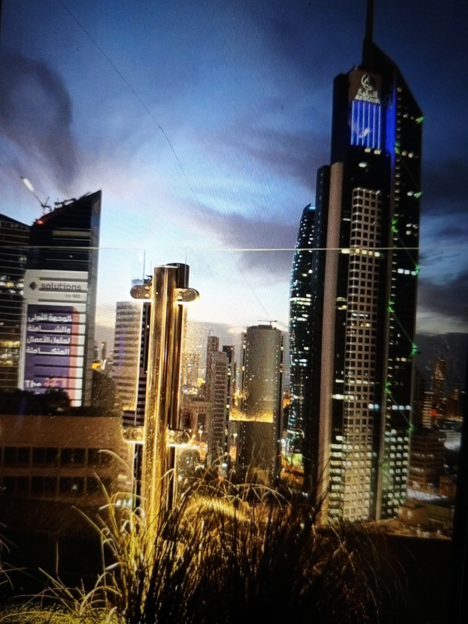 The image shows a cityscape at dusk, with several skyscrapers illuminated by lights. In the foreground, tall, dry grasses are silhouetted against the city lights. The sky is a deep blue, transitioning to lighter shades near the horizon, suggesting the end of the day.
The most prominent building in the center is a very tall skyscraper with a dark facade. It has a distinctive logo at the top and vertical blue lights running down a section of its side. To its left is another skyscraper with a more angular design. A large sign on this building displays "solutions" in white text on a dark background. Below this, a purple banner has several lines of Arabic text, followed by "The 471".
Other buildings of varying heights and designs are visible in the background, some with grid-like window patterns and others with scattered lights. The overall impression is one of a modern, vibrant urban environment at twilight. The presence of the dry grass in the foreground suggests an outdoor viewpoint, possibly from a balcony or a higher elevation overlooking the city. There is no discernible activity or event taking place; it is a static scene of urban architecture. The lighting and the time of day create a somewhat dramatic and sophisticated atmosphere.