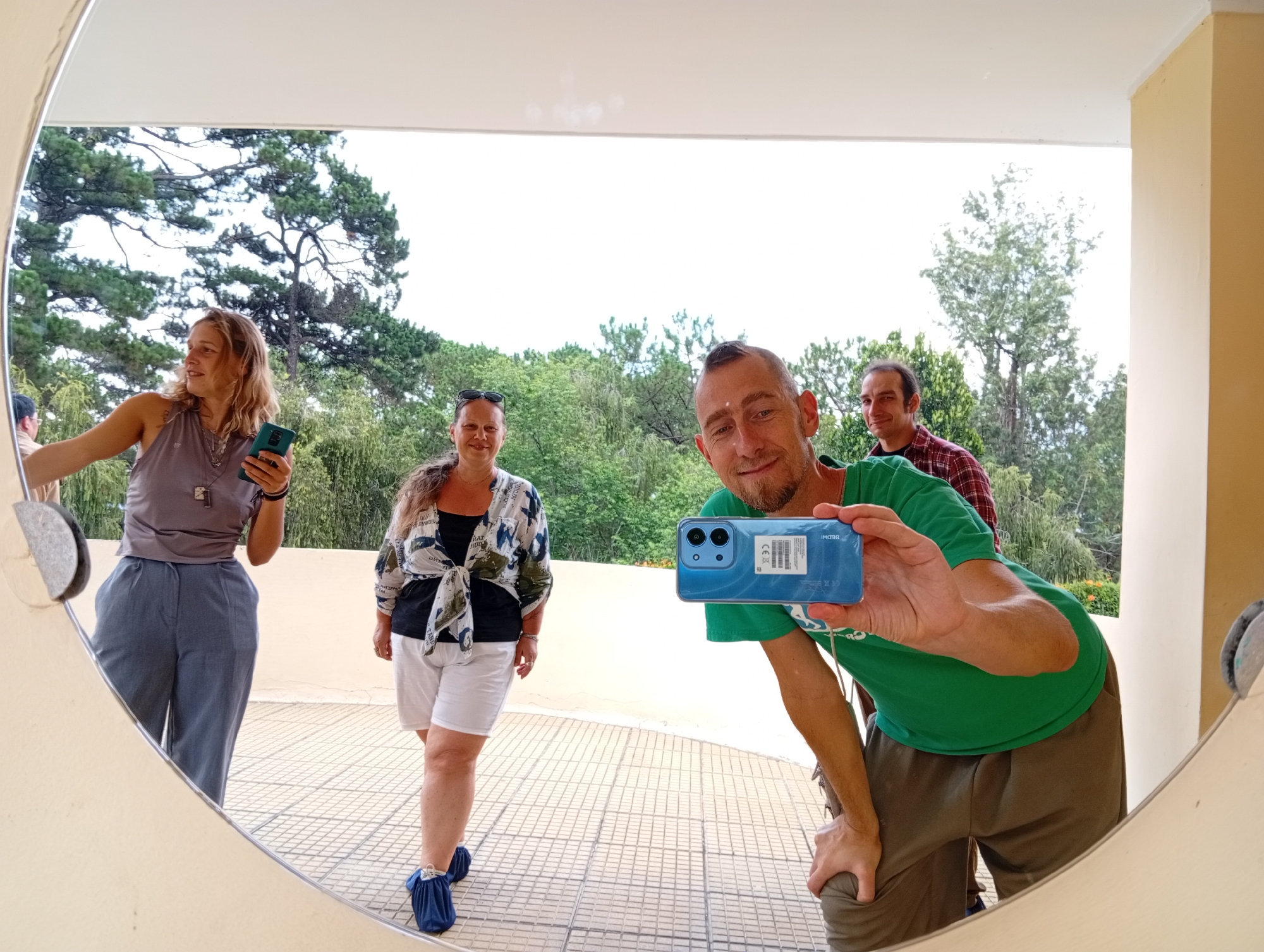 A medium indoor shot depicts four individuals reflected in a large, round mirror against an outdoor backdrop, possibly in Da Lat, Vietnam, based on architectural cues and landscape features.
On the left, a fair-skinned woman with blonde hair, wearing a gray top and high-waisted blue pants, stands holding a blue smartphone in her right hand, seemingly interacting with the mirror's surface. A middle-aged woman stands slightly behind her and to the right. She has dark hair, sunglasses perched on her head, and wears a black top beneath a light-colored floral-patterned shirt tied at the waist, paired with white shorts and blue shoe covers. Further to the right, a man in a green t-shirt, tan pants, and closely cropped hair, leans slightly towards the mirror, holding a blue smartphone horizontally in front of him, likely capturing the reflected scene. Behind him and to the left, a fourth individual, also male, with darker hair, stands wearing a red and black plaid shirt. The mirror frame is a light beige or off-white color.
The outdoor background is visible through the mirror's reflection and shows a tiled terrace or balcony and a lush landscape featuring a variety of trees, suggesting a mountainous or elevated terrain. Above, a white ceiling is partially visible above the reflected scene.