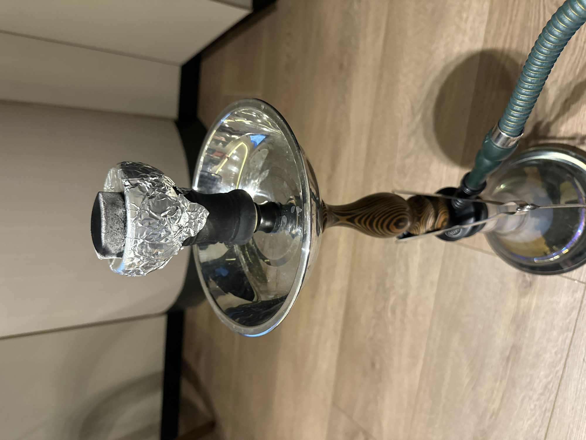 The image depicts a hookah with a silver bowl wrapped in aluminum foil, a wooden patterned stem, and a green hose, placed on a light-colored wooden surface. A white cabinet is partially visible in the upper left corner. The location appears to be indoors, potentially a home or establishment, but the image doesn't provide enough information to place the scene in Mexico City, Mexico.