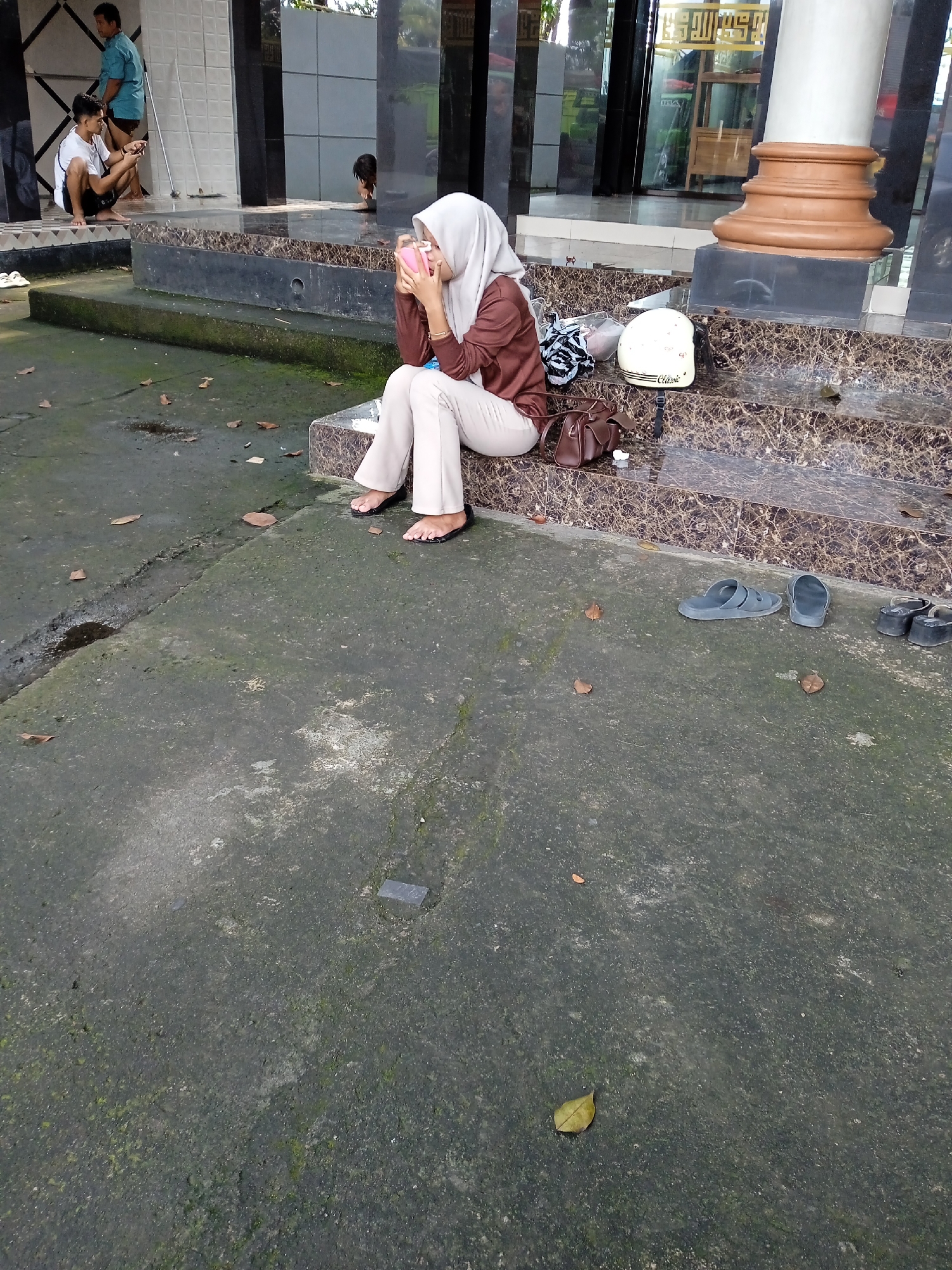 The photo shows a woman wearing a hijab and brown shirt, seated on a marble staircase. She is holding a pink object to her face. Beside her are a white helmet, a patterned bag, and a brown purse. Various pairs of sandals are near the bottom step. The concrete ground in front of the staircase has a mottled, greenish appearance, with scattered leaves. In the background, two men are visible near a building with reflective windows. One of the men is sitting, and the other is standing. The location is Wonoketingal, Indonesia.