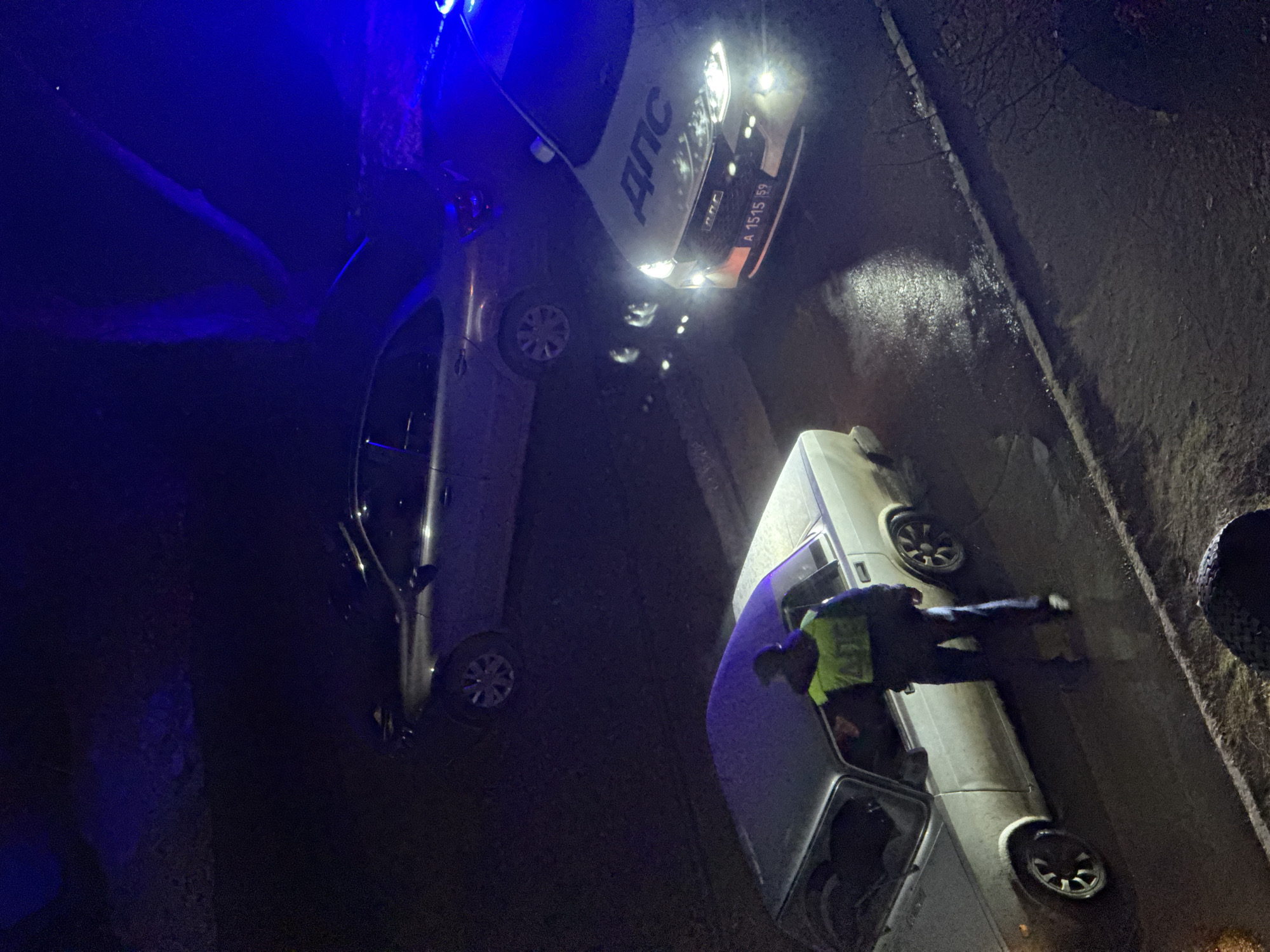 At night on a city street in Chaikovsky, Russia, two cars are present, along with a person who appears to be law enforcement. One car is light-colored with an open door, while the other is white with a clearly visible plate number A 1515 59. The law enforcement officer is standing near the open door of the light-colored car. Blue lights suggest the presence of law enforcement activity. The street appears wet.