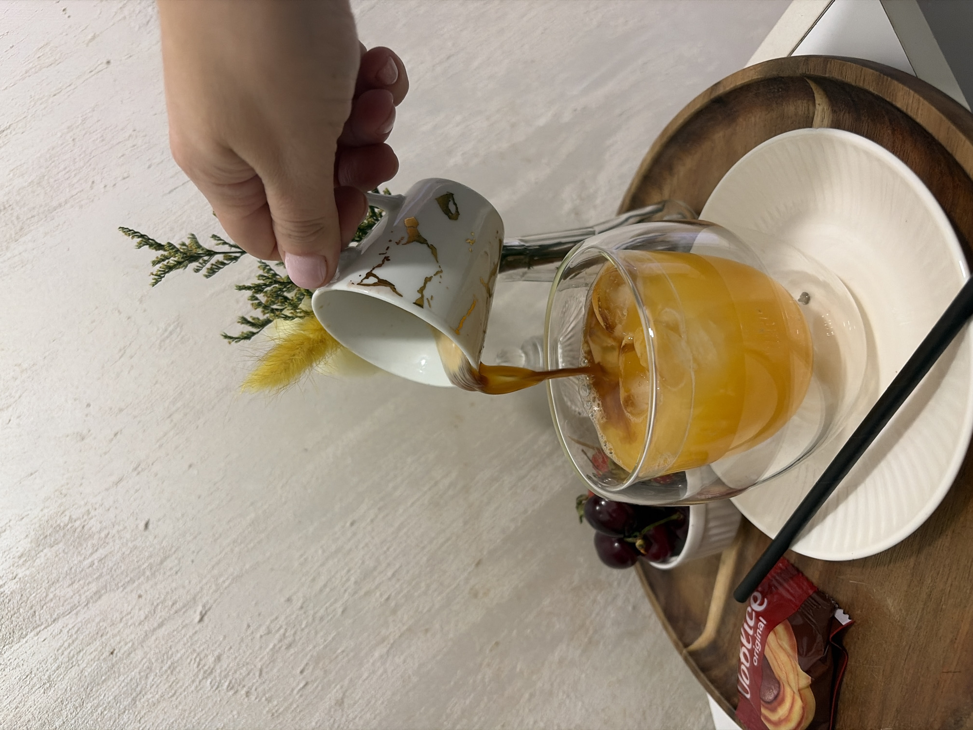 The image shows a hand pouring coffee from a white and gold mug into a glass filled with orange liquid and ice, likely a cocktail or juice mix. The drink is placed on a wooden tray with a white plate, a small bowl of cherries, a black straw, and a Voortice original chocolate biscuit wrapper. There's a sprig of green and yellow dried flowers near the mug. The background is a textured, off-white surface. The precise location in Budva, Montenegro, cannot be determined without additional information.