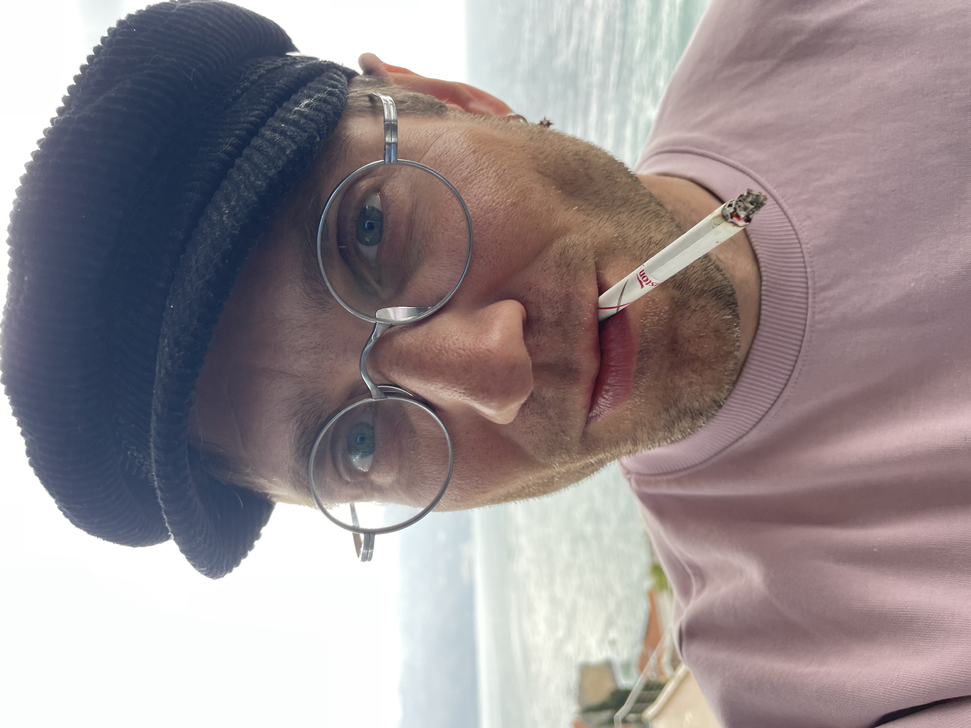 A male individual is depicted in a close-up, chest-up shot, standing outdoors with the sea and distant landforms of Budva, Montenegro, visible behind him. He wears a dark corduroy newsboy cap and round, metal-framed glasses. He has light facial stubble and a silver hoop earring in his right ear. He is dressed in a light pink crew-neck sweatshirt. A lit cigarette, partially labeled "Winston," is held between his lips, featuring a visible ember and ash at its tip. He looks directly at the viewer. The background shows calm, light blue-green sea water extending to a hazy horizon with distant landmasses, characteristic of the Budva coastline. Residential rooftops with orange tiles and a light-colored railing are visible in the lower-left background. The sky is overcast, providing soft, diffused lighting.