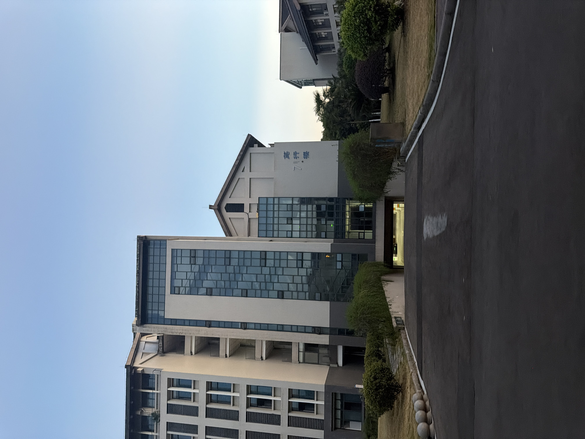The image depicts a street view in front of a modern building complex, likely an educational institution, in Jiangjin, China. The architecture features a tall, multi-story building with a facade of alternating grey panels and blue glass windows. Adjacent to it is another building with a pitched roof and a similar modern design. The ground is paved with asphalt, forming a sloping driveway that leads towards an underground parking entrance. Landscaping includes manicured bushes and trees on either side of the driveway and surrounding the buildings, with a clear blue sky overhead. A sign with blue Chinese characters is visible on the light-colored wall of one of the buildings, reading "7# Building". There are no people visible in the image.