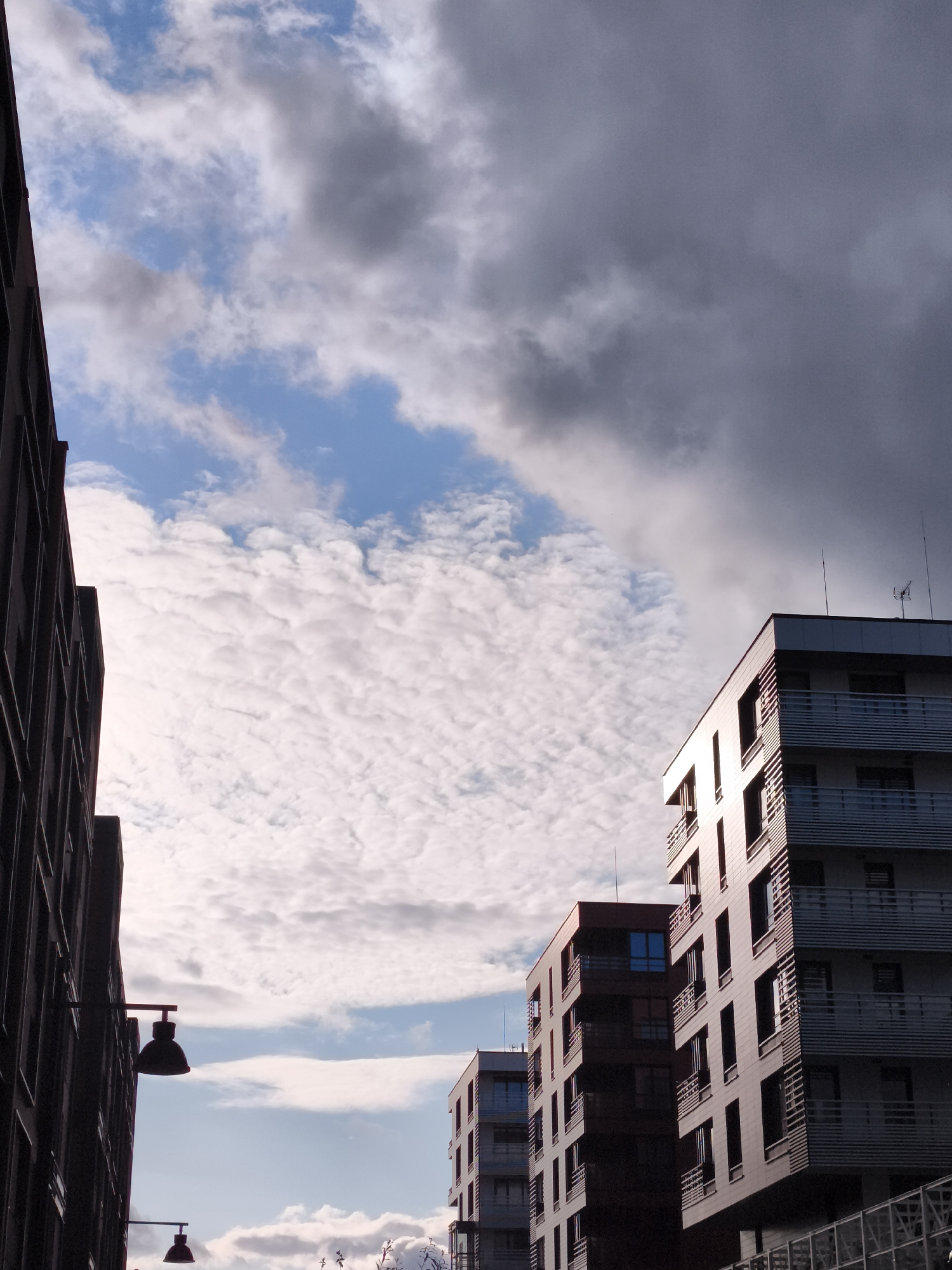 This is a low-angle, vertical shot looking up at modern apartment buildings against a dramatic sky in the city of Gdańsk, Poland. The sky is a mix of bright blue with fluffy white clouds and ominous, dark grey storm clouds gathering from the right. The buildings are contemporary in design with multiple stories, balconies with horizontal railings, and numerous windows. On the left, a dark, tall building with visible window frames is partially visible, suggesting it is closer to the camera. To the right, two lighter-colored apartment buildings are prominent, one slightly taller and more angular than the other. The tallest building on the right has an antenna and what appears to be a satellite dish on its roof. Two dark, decorative lamps are attached to the side of the building on the left. The overall impression is of an urban setting with contrasting architectural styles and a dynamic weather pattern.