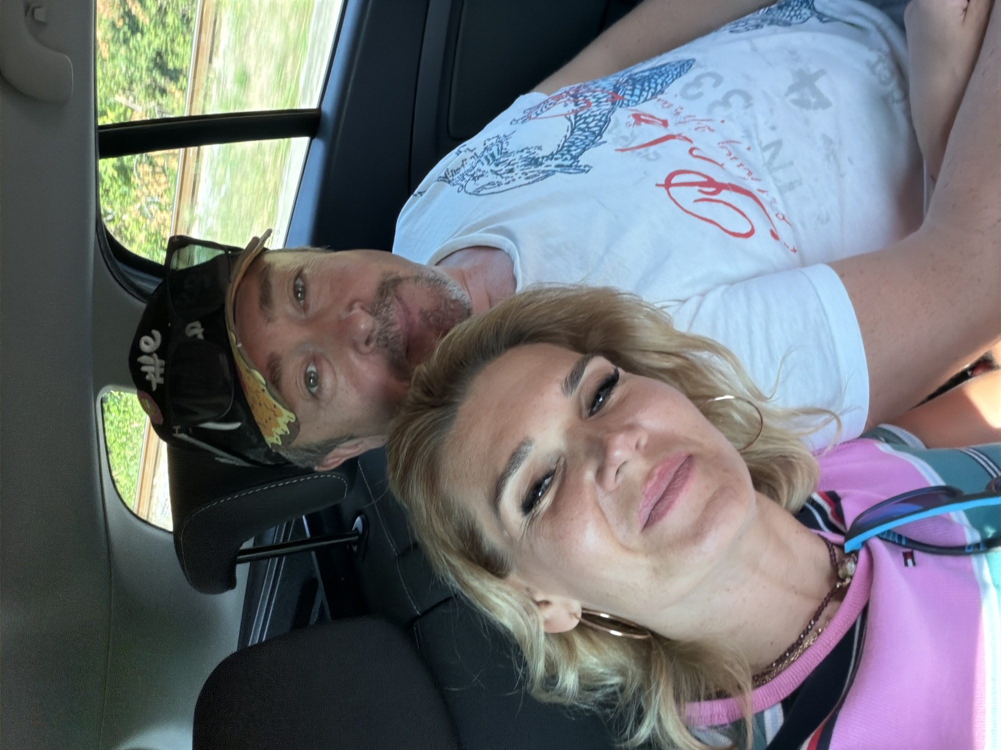 A man and a woman are in the back seat of a car, taking a selfie. The woman is in the foreground, her face tilted towards the camera. She has blonde hair, hoop earrings, and is wearing a pink and striped shirt. The man is behind her, his head slightly turned to the side. He is wearing a cap and a white t-shirt with a graphic print. The car interior is visible, including the headrests and the window, through which greenery and what appears to be train tracks can be seen. The location is the city of Bistrice, Montenegro.