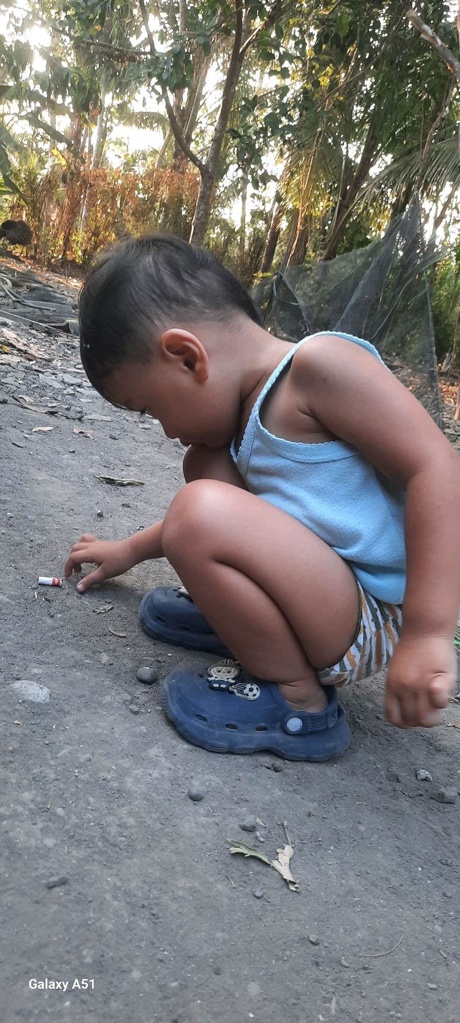 A young child, possibly a toddler, is crouching on a dirt ground in what appears to be a rural setting. The child is wearing a light blue tank top and shorts with a striped pattern. They have dark hair and are looking down at something on the ground with their right hand reaching out. In front of the child, there is a small, red and white cylindrical object, possibly a cigarette butt. The ground is rough and uneven, with some small stones and dry leaves scattered around. In the background, there are trees with dense foliage and some sunlight filtering through, suggesting an outdoor environment. A dark net-like structure is visible behind the child. The lower right corner of the image displays "Galaxy A51".