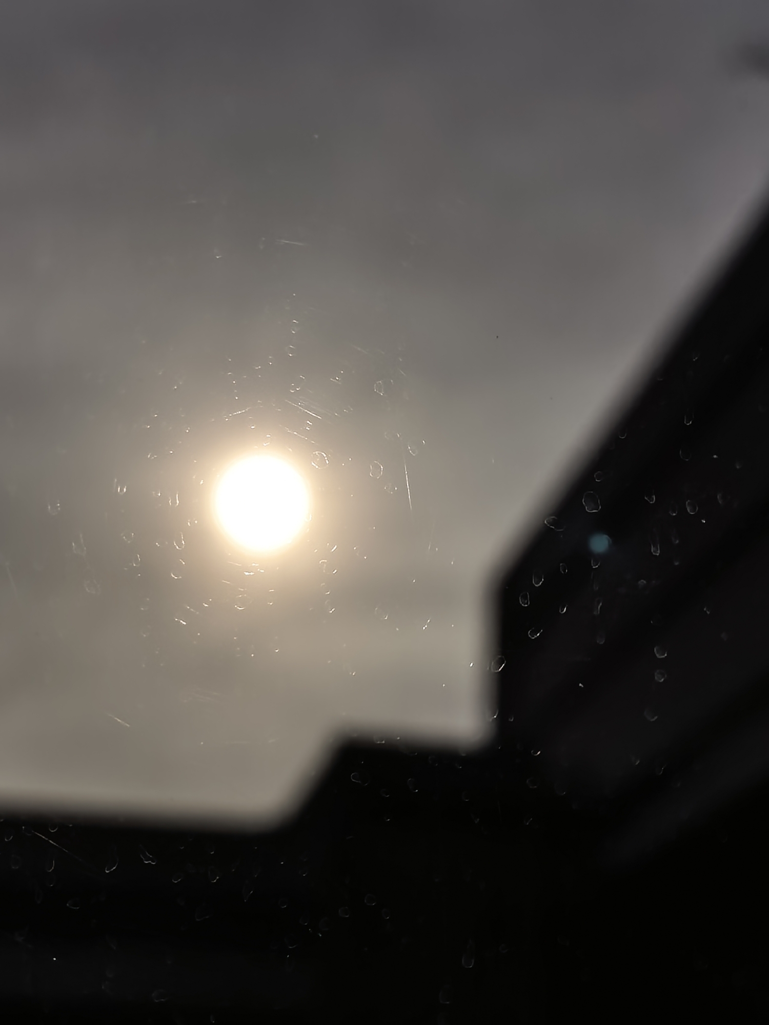 The image captures a bright, circular light source, likely the sun, centered slightly to the left, emanating a strong glow through what appears to be a wet or dirty glass surface. Numerous water droplets and some streaks are visible on this surface, distorting the light and the background. The sky behind the light source is a hazy, muted grey, suggesting an overcast day or atmospheric conditions affecting visibility.
To the right and bottom of the frame, there are blurred, dark, angular shapes that resemble the silhouette of a building or a window frame, creating a stark contrast against the bright light and grey sky. A small, faint blue lens flare is also visible among the water droplets in the lower right quadrant. The overall impression is that of looking out a window or through glass on a day with rain or condensation, with a strong light source partially obscured by the elements and surrounding structures. There is no visible text.