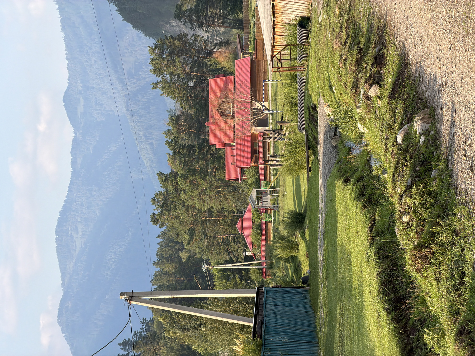The image depicts a rural scene in the village of Askat, Russia, bathed in the soft light of late afternoon. In the foreground, a gravel road curves to the right, bordered by a grassy embankment with a small, shallow stream running through it. A bright green corrugated fence is visible on the left, next to a teal-colored metal structure.
Further back, several wooden houses with vibrant red tiled roofs are nestled among lush green trees. A children's playhouse with a slide stands on a manicured lawn. In the middle ground, a dense forest of tall pine trees stretches across the landscape. In the distance, a series of majestic, mist-covered mountains form a breathtaking backdrop, their peaks softened by atmospheric haze. A tall utility pole with electrical wires runs through the scene, indicating the presence of infrastructure in this remote location. The overall impression is one of a peaceful, idyllic rural setting.