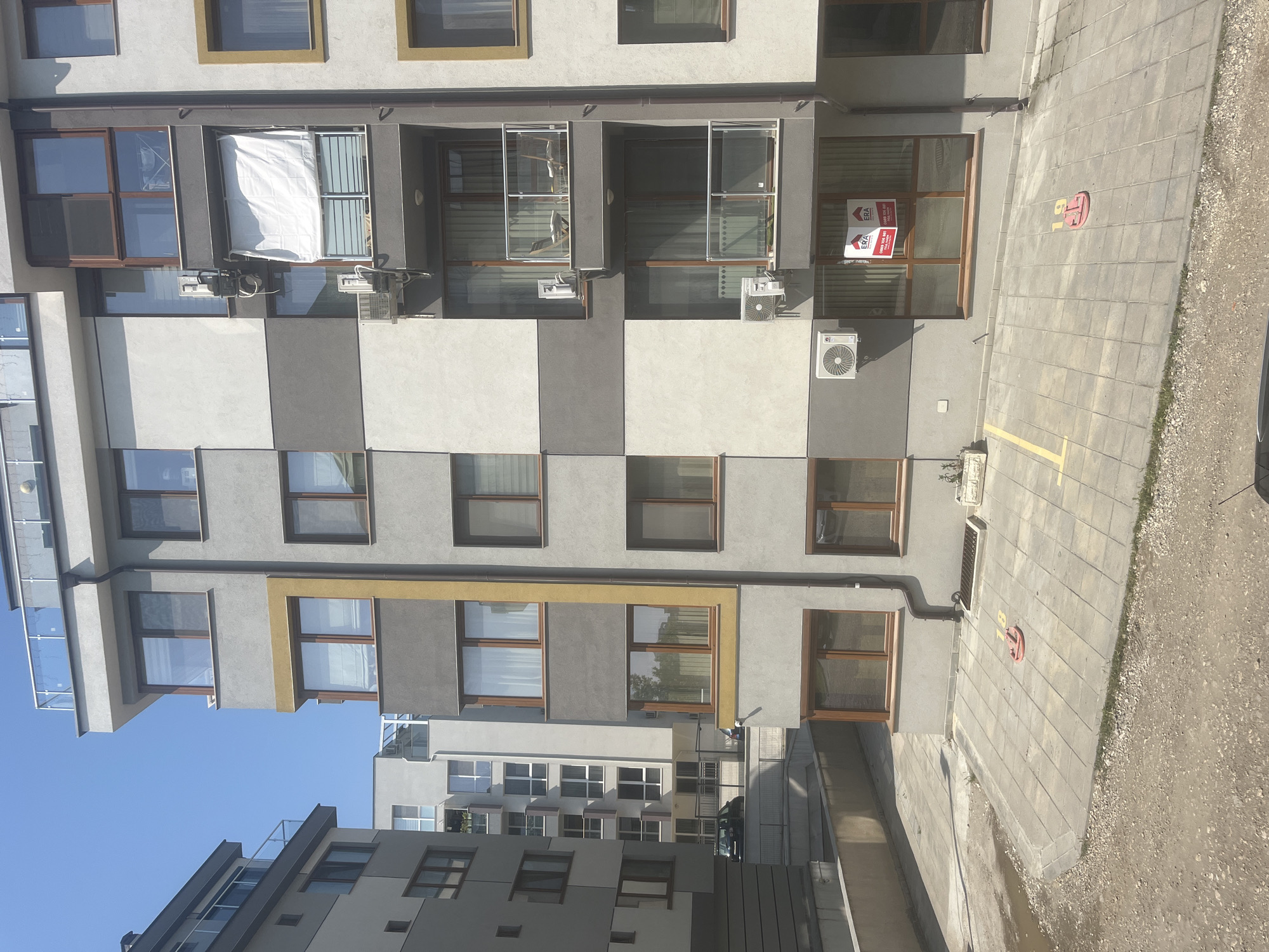 The image depicts a modern, multi-story building facade under a clear, bright blue sky, suggesting a sunny day. The building features a contemporary design with alternating light gray and dark gray rectangular panels. Some window frames are brown, while others are highlighted with a golden-yellow border.
On one of the upper-level windows, a large white sheet or piece of fabric is draped over a railing, possibly drying. Below this, several outdoor air conditioning units are mounted on the exterior walls.
At the ground level, there's a paved area with gray square tiles, featuring yellow painted lines, likely for parking. Adjacent to the pavement on the right is a rougher patch of gravel or dirt. A prominent red object, possibly a parking barrier or marker, with the number "18" in yellow, rests on the paved surface.
One of the ground-floor windows displays a red and white real estate sign. Visible text on the sign includes "ERA" (the company logo), "FOR RENT" in English, and "ПОД НАЕМ" (For Rent) and "ПРОДАВА" (For Sale) in Bulgarian, along with a partial phone number starting with "0898".
In the background, other similar modern buildings are visible, indicating an urban or residential development. No people are present, and no discernible activity is taking place beyond the static display of the buildings and the real estate sign. The overall scene is well-lit by natural daylight.