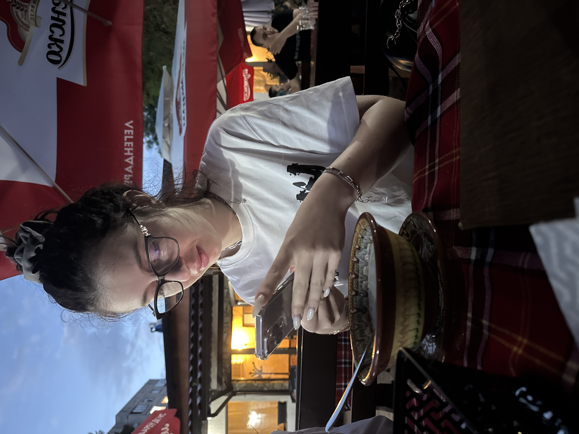 The image depicts a woman with dark hair pulled back in a scrunchie, wearing glasses and a white t-shirt, sitting at a table and looking at her smartphone. A bowl is positioned on the table to her right. The table is covered with a red and black plaid tablecloth. In the background, there is a red banner with the word "VELEHIV" printed on it and an umbrella. Other individuals are visible in the background. The location is suggested to be Sofia, Bulgaria, based on the flags and the architecture. The scene appears to be an outdoor dining area, likely a restaurant or café.
