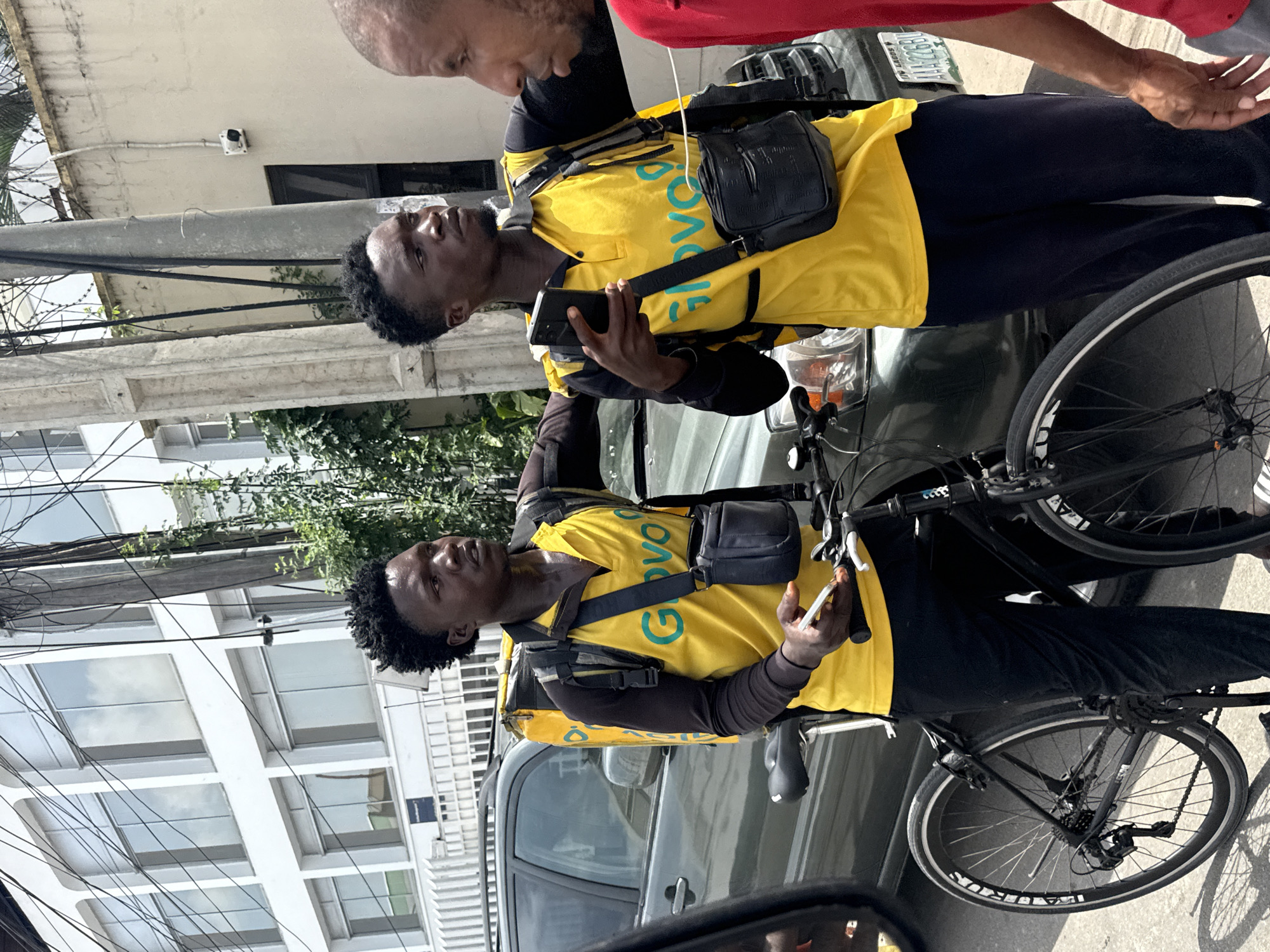 Two individuals in Lagos, Nigeria, are engaged in a conversation near a dark gray vehicle. Both individuals are wearing yellow Glovo shirts. One of the two individuals holds a cell phone. Both have black satchels, one cross-body and the other on his back. There is a bicycle standing between the front of the vehicle and the two individuals.