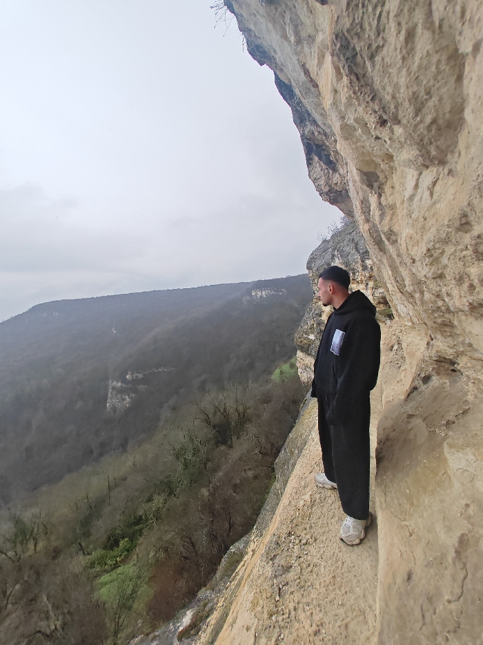 One male subject is depicted in profile, standing on a narrow, light-colored rock ledge, approximately one meter wide, adjacent to a textured limestone cliff face that dominates the right portion of the image. The subject, a young adult, wears a black hooded sweatshirt with a light rectangular emblem on the left chest, black trousers, and light-colored athletic shoes. His head is turned left, observing a vast, deep forested valley containing predominantly bare deciduous trees and sporadic green ground cover, with distant hills receding into an overcast grey sky. The scene is situated on the Shumen Plateau, within the municipality of Shumen, Bulgaria.