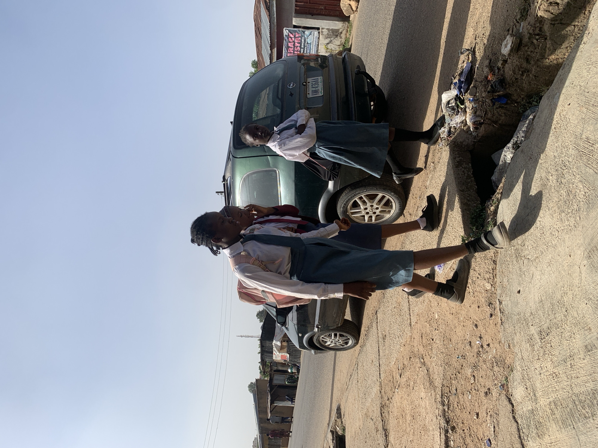Three children in school uniforms stand beside a parked green minivan on a street in Gura Top, Nigeria. The street is unpaved in areas and borders a concrete sidewalk with a deep, trash-filled gutter. Buildings are visible in the background. The sky is clear and bright. One child is leaning against the car, while another appears to be speaking to someone out of frame.