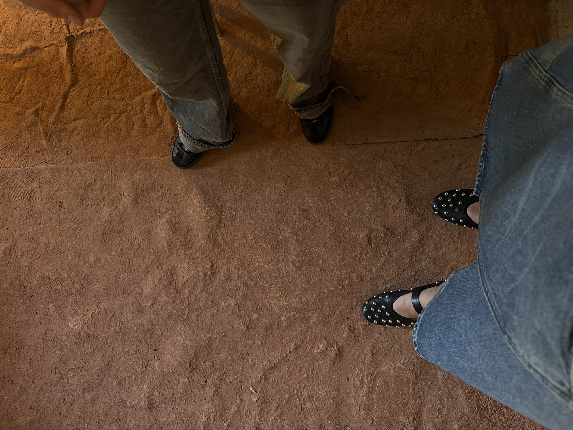 The image shows a view of three pairs of legs and shoes. The legs are wearing jeans of different shades. The shoes on the left and center are black, casual shoes. The shoes on the right are black flats with studs. The background is a reddish-brown stone-like surface. The environment suggests an outdoor setting or a place with a rustic interior. There is no information in the image to verify the location in Milan, Italy.
