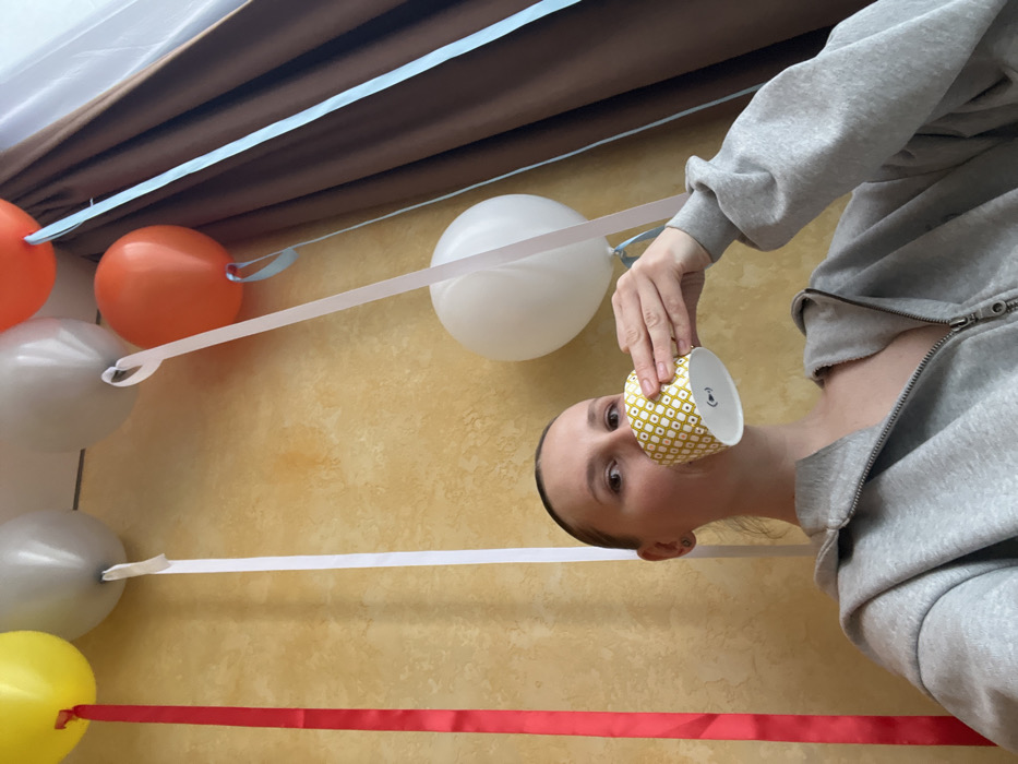 The image displays an indoor scene located in the city of Khimki, Russia. A fair-skinned individual, appearing female, with light brown hair pulled back, is positioned in the foreground, looking directly at the camera. She is wearing a grey zippered hooded sweatshirt, with the zipper open to expose the upper chest area. Her right hand holds a yellow and white geometrically patterned disposable cup to her mouth, from which she is in the process of drinking. The background features a textured, light yellow wall to the left and center, and a dark brown curtain on the right side. Several helium-filled balloons are visible in the upper portion of the frame, near the ceiling. These include a yellow balloon with a red ribbon, two grey balloons with white ribbons, two orange balloons with light blue ribbons, and a partially visible white balloon also with a light blue ribbon. Ribbons of red, white, and light blue colors hang vertically from the balloons.