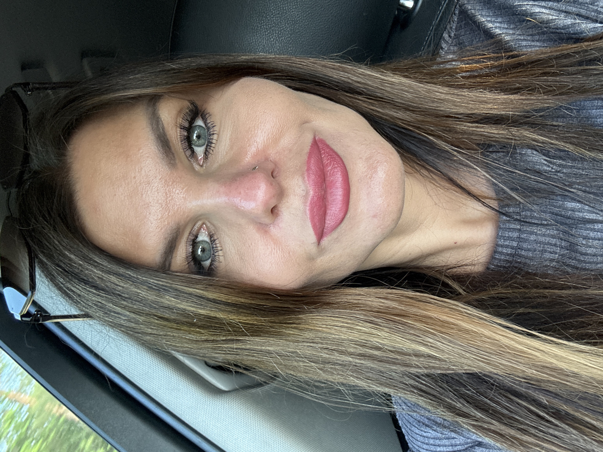 A close-up, front-facing selfie shows a woman in a car. She has long, layered brown hair with blonde highlights, parted in the middle, and appears to be wearing a grey ribbed top. Her makeup includes defined eyebrows, dramatic false eyelashes, and pink lipstick. She is looking directly at the camera with a neutral expression. Sunglasses are perched on top of her head, and a small nose ring is visible. The background of the car interior is out of focus, with a glimpse of green foliage through the window on the left, suggesting an outdoor setting. The image does not provide specific details that would allow for pinpointing a location within Chaikovsky, Russia.